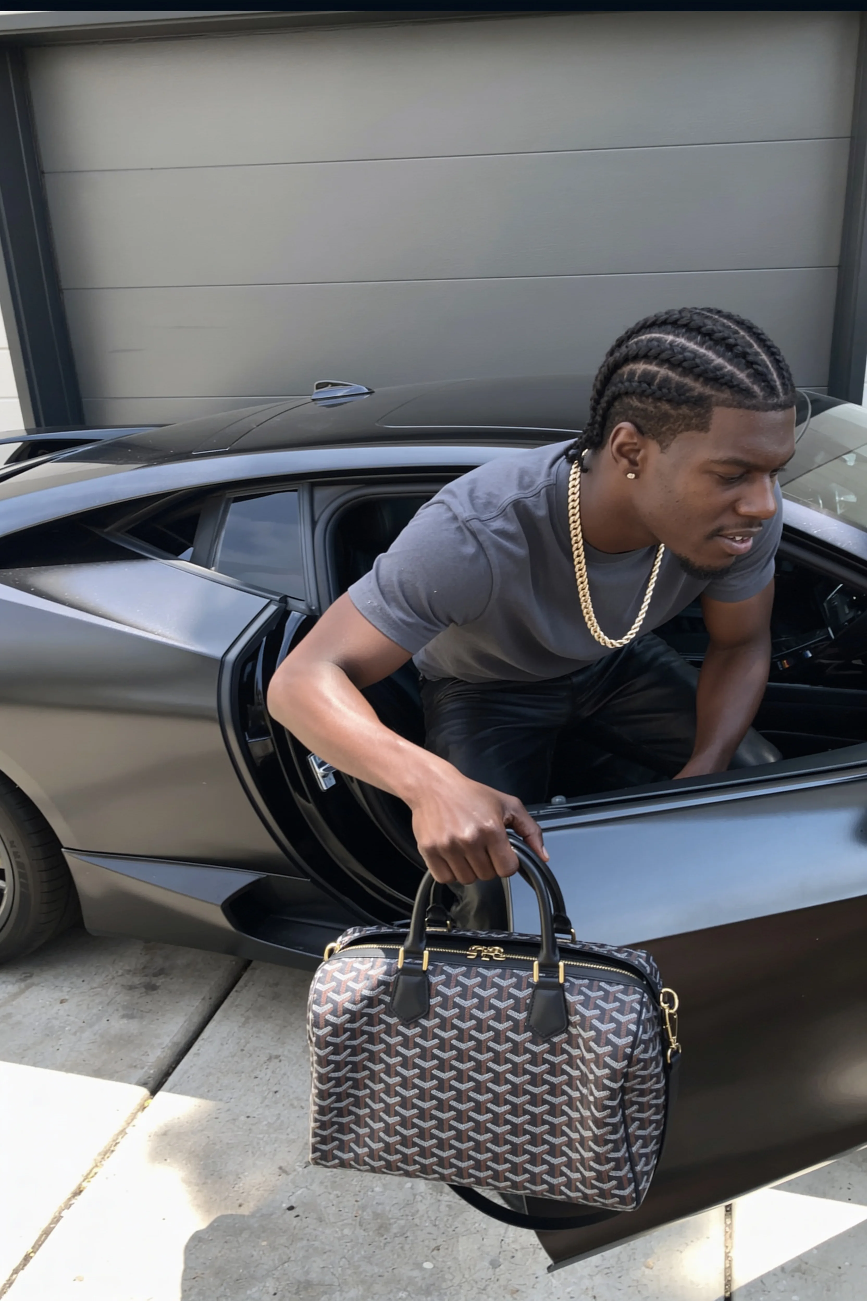 A person with braided hair and a gold chain is kneeling next to an open car door, holding a patterned handbag.