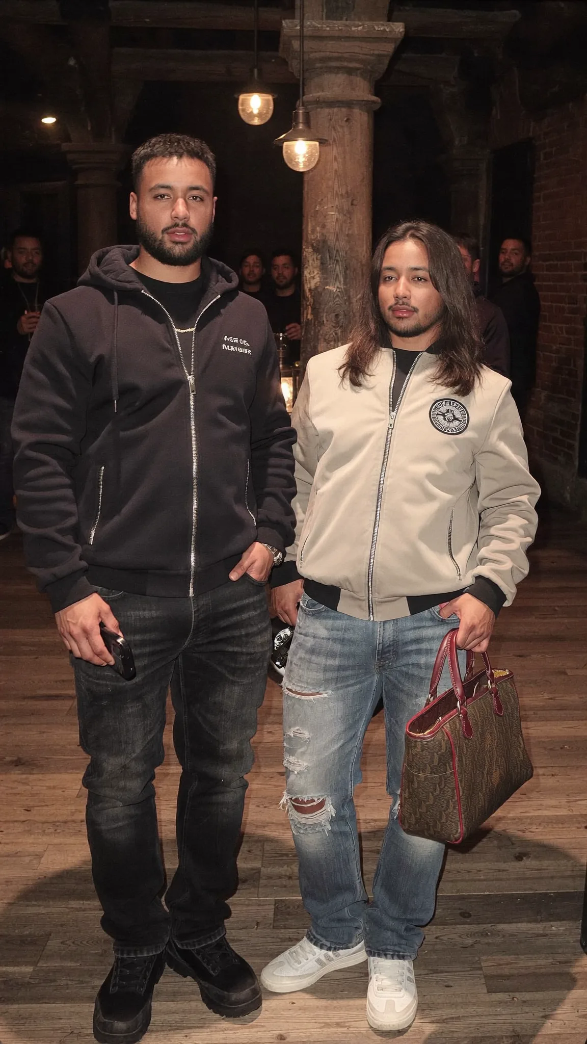 Two men stand together in a casual setting, one holding a handbag.