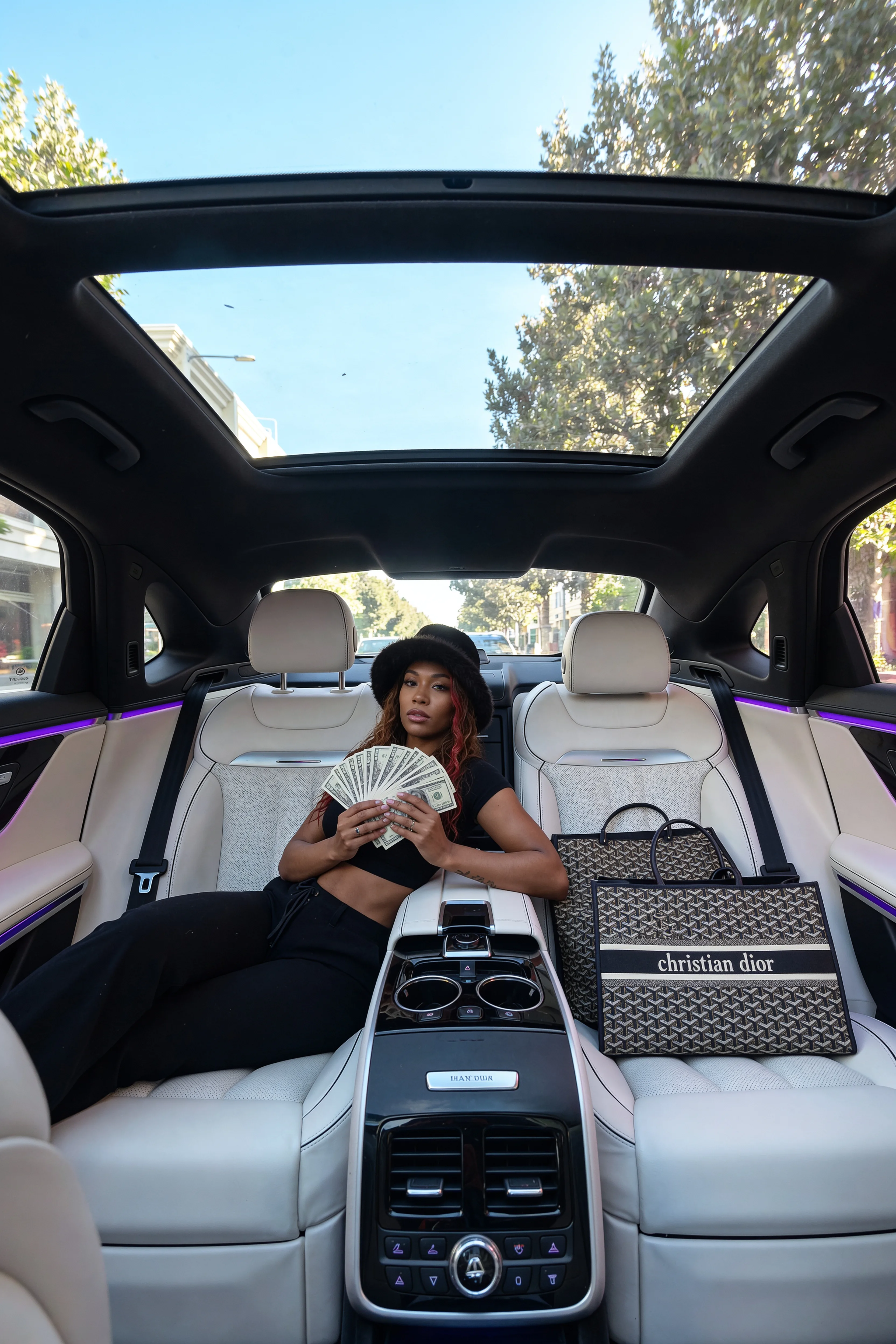 A woman with platinum blonde hair is seated in a luxury car, holding money and wearing casual attire.