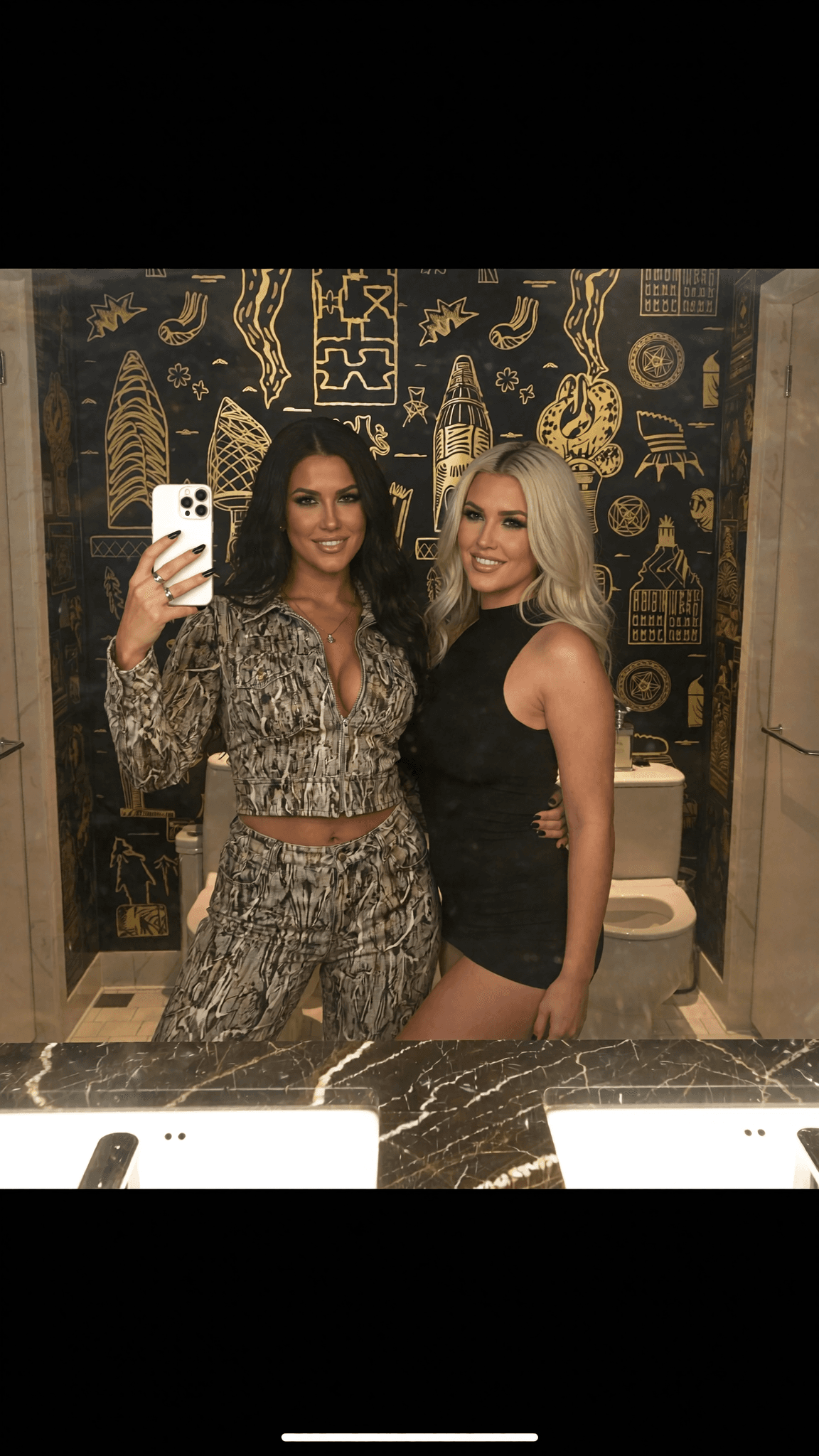 Two women pose in a stylish bathroom, one wearing a dress and the other dressed casually.