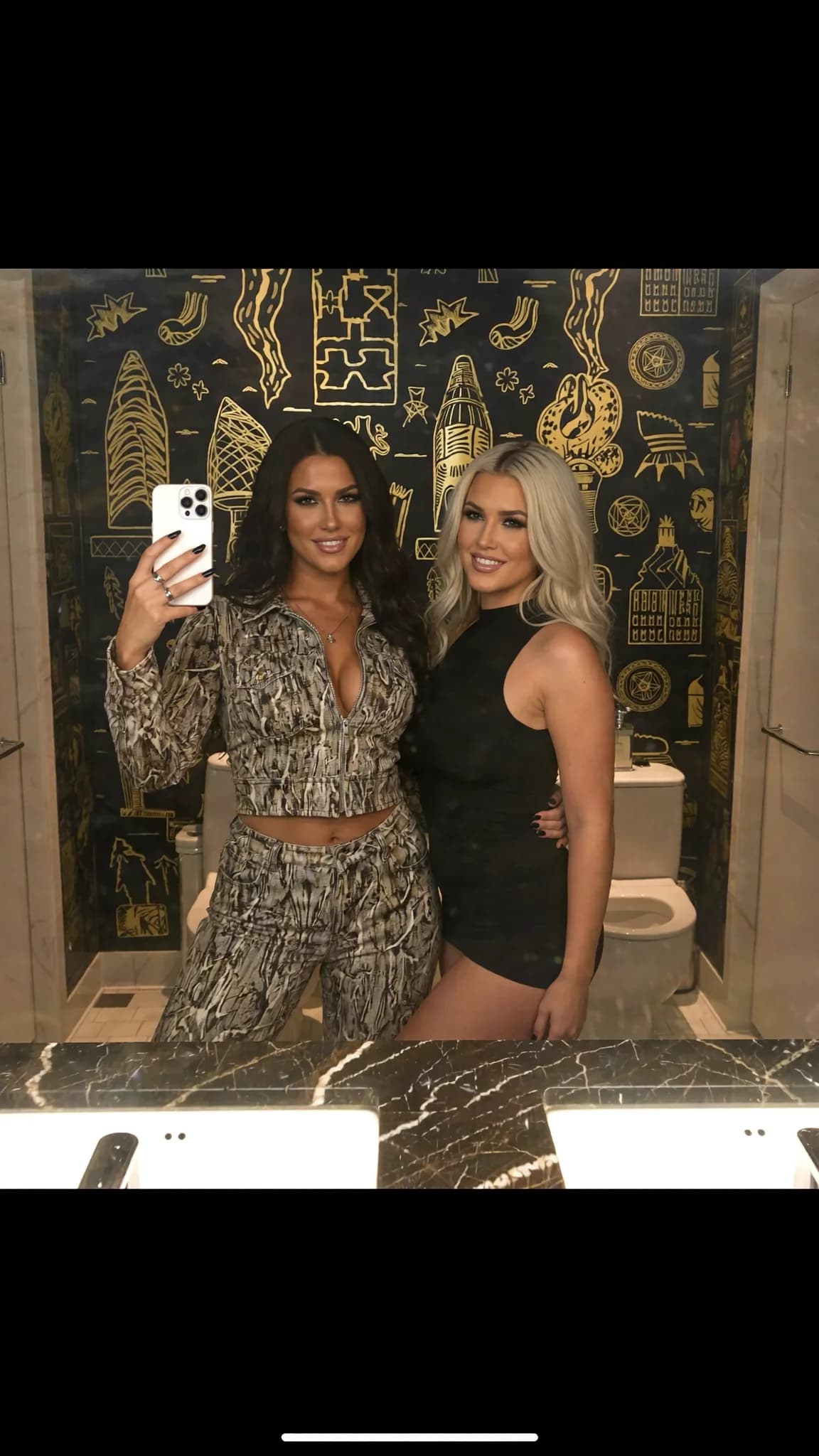 Two women pose in a stylish bathroom, one wearing a dress and the other dressed casually.