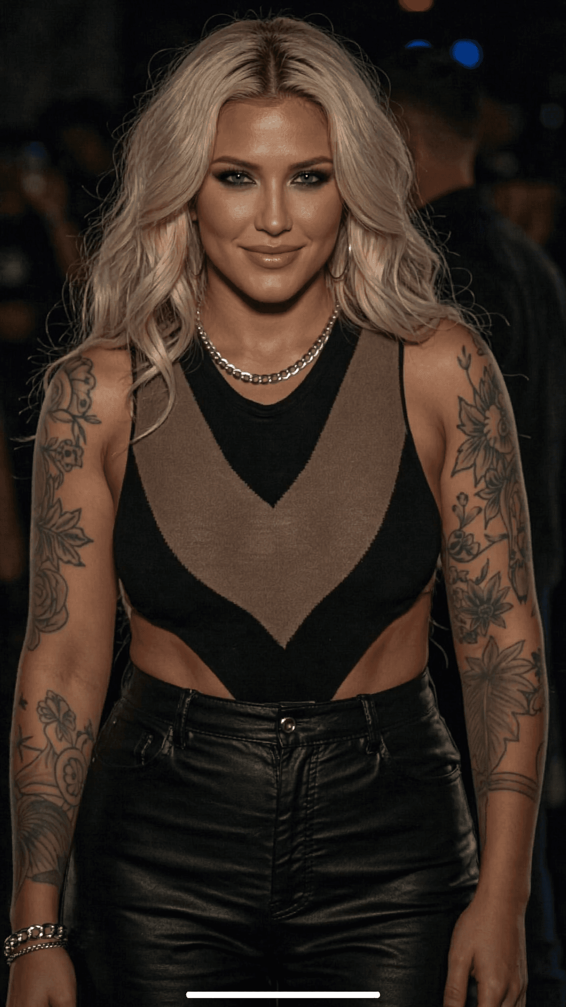 A woman with platinum blonde hair and tattoos stands confidently in an outdoor setting, wearing a casual outfit.