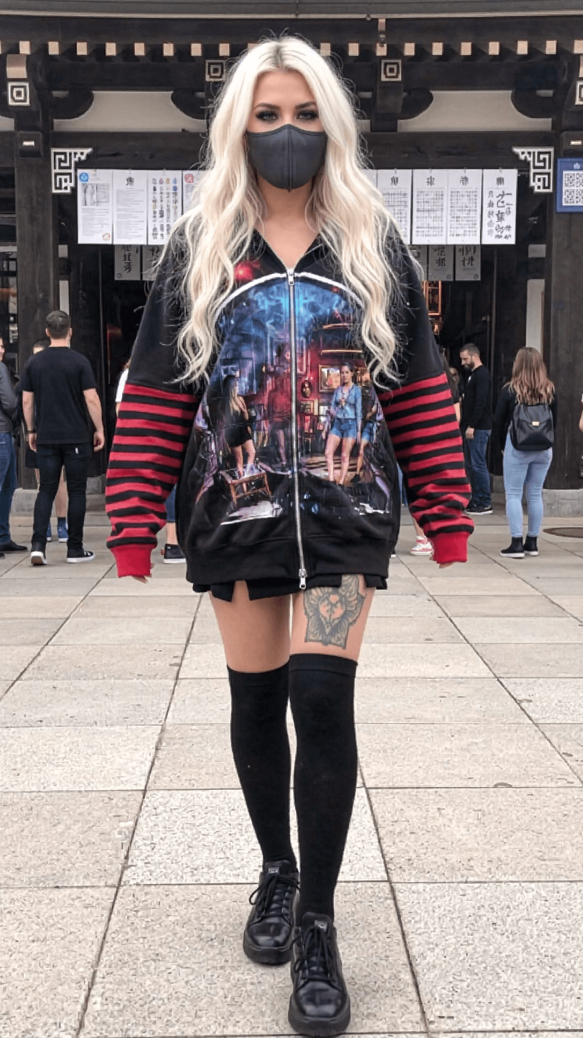 A woman with platinum blonde hair and a black mask stands confidently in an outdoor setting, wearing a graphic hoodie and thigh-high socks.