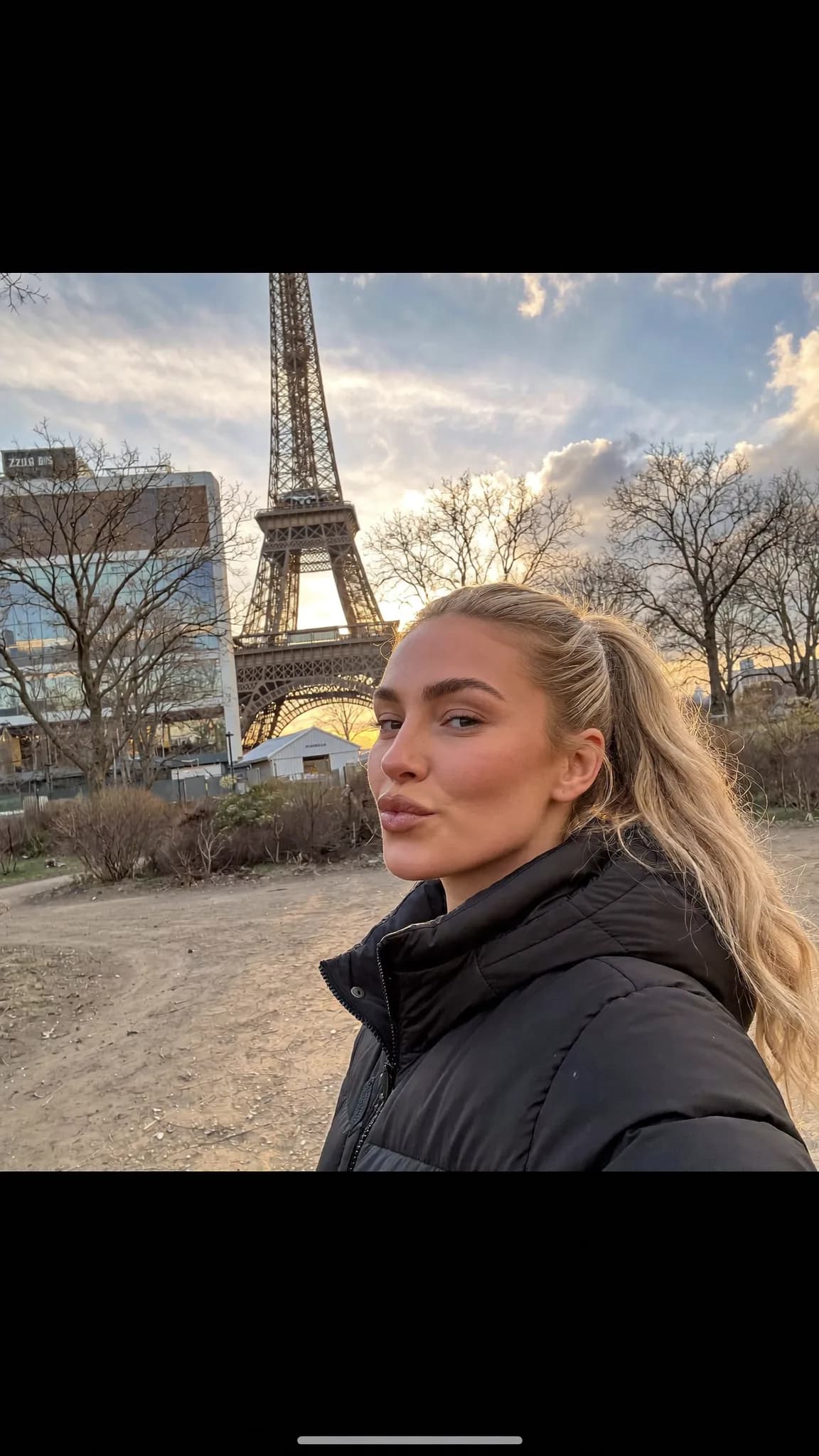 A woman with blonde hair poses in front of the Eiffel Tower during sunset.