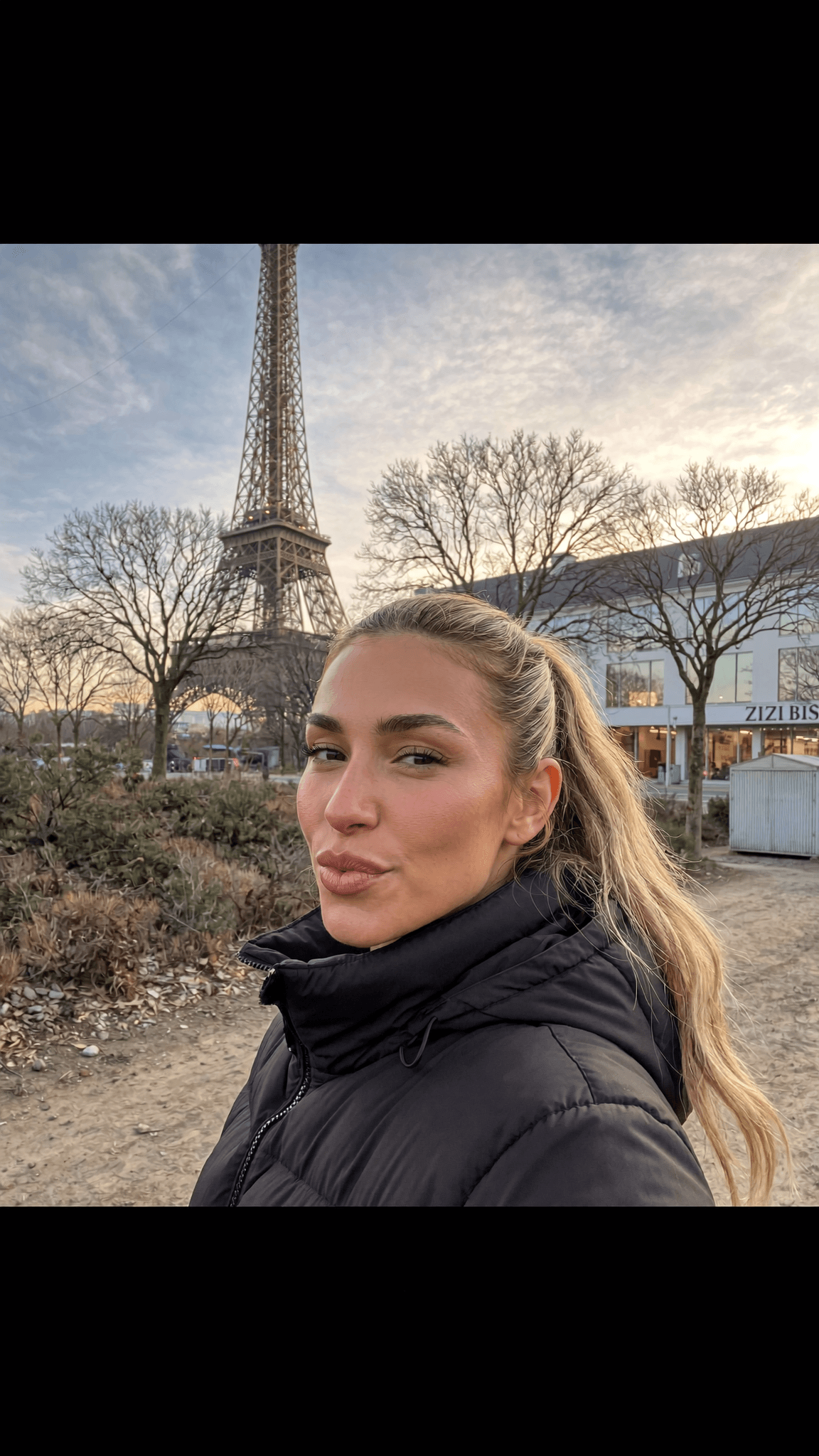 A woman with blonde hair poses in front of the Eiffel Tower wearing casual clothing.