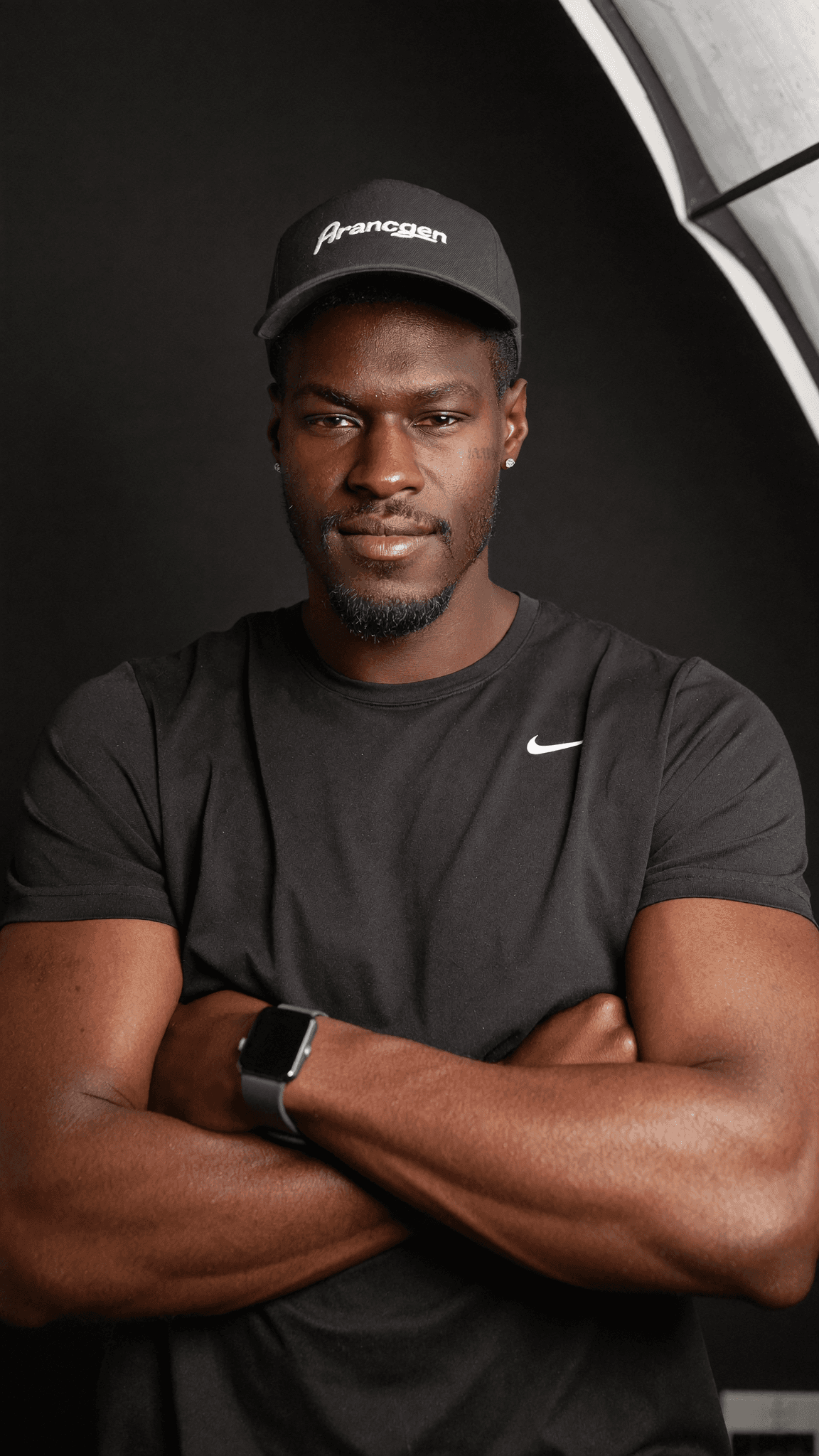 A man in a black Nike shirt and cap poses confidently with arms crossed against a dark background.