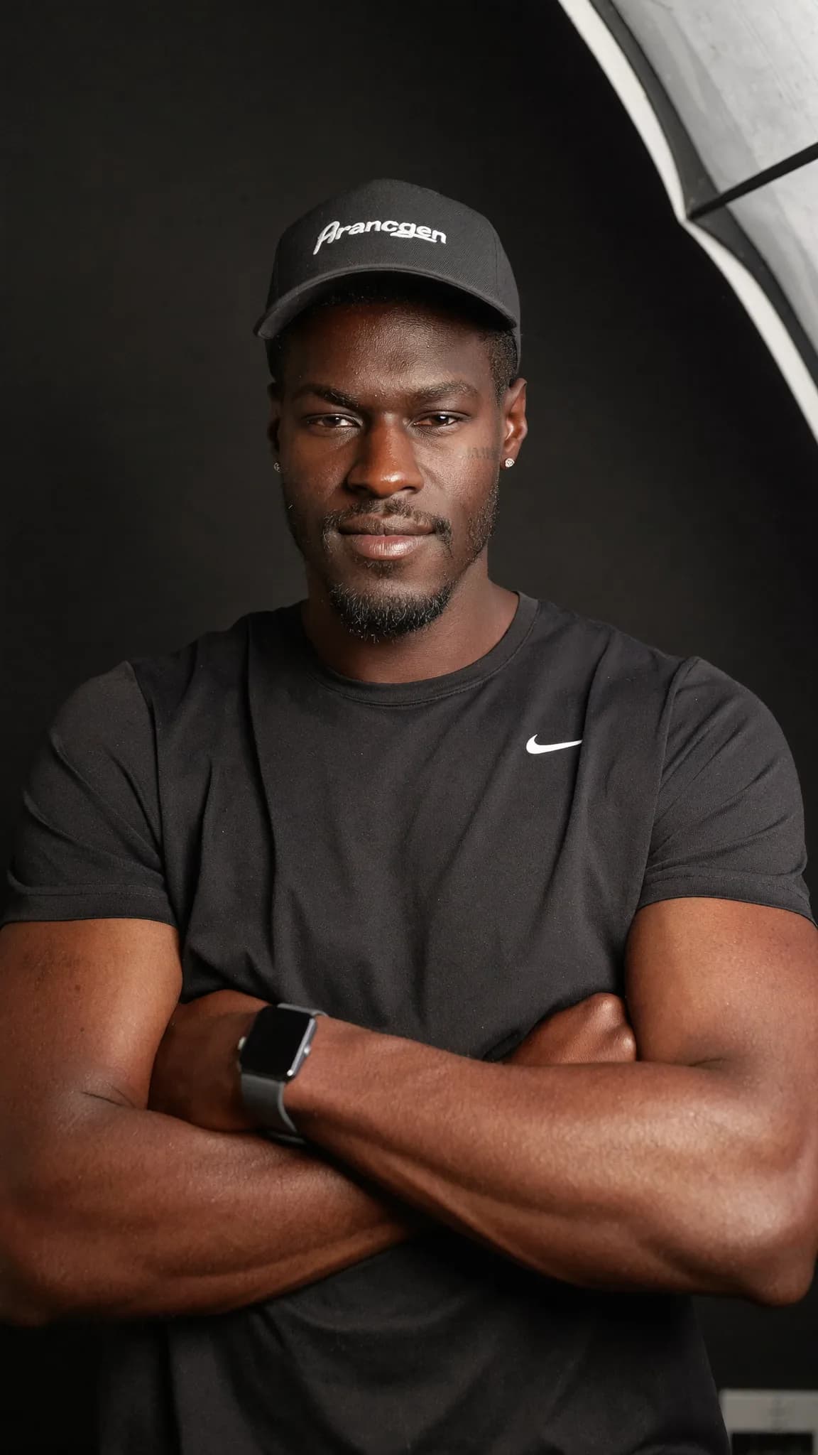 A man in a black Nike shirt and cap poses confidently with arms crossed against a dark background.