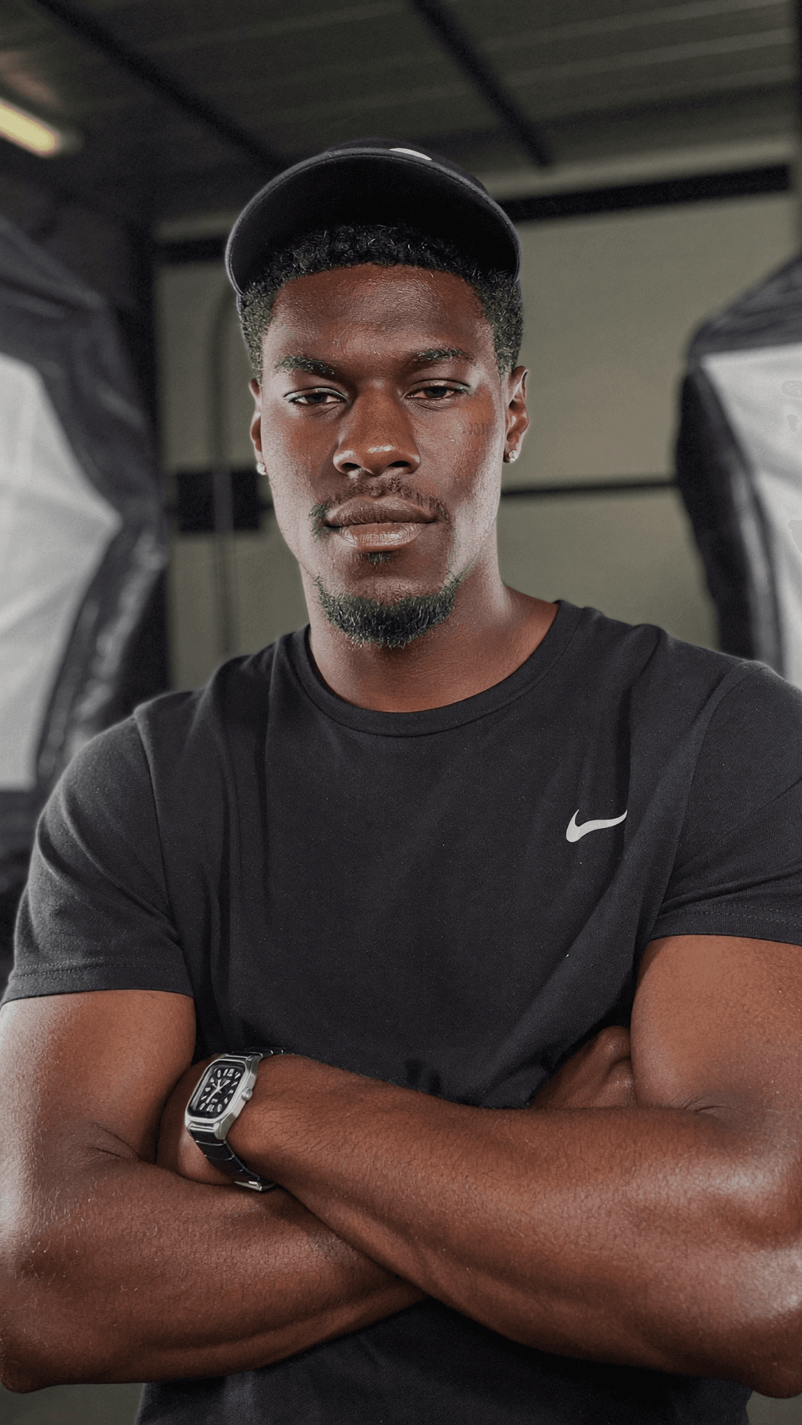 A man in a black Nike shirt and cap stands confidently with arms crossed in a studio setting.