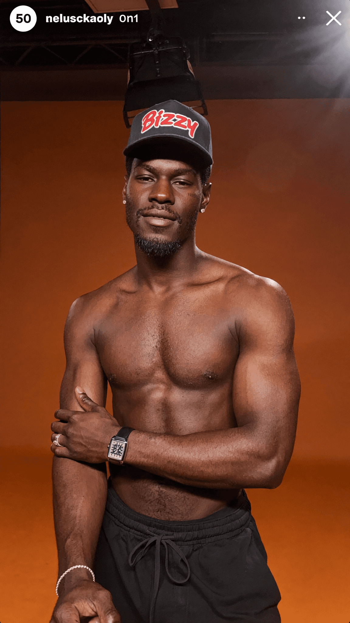A shirtless man wearing a cap and black shorts poses confidently in a studio setting.