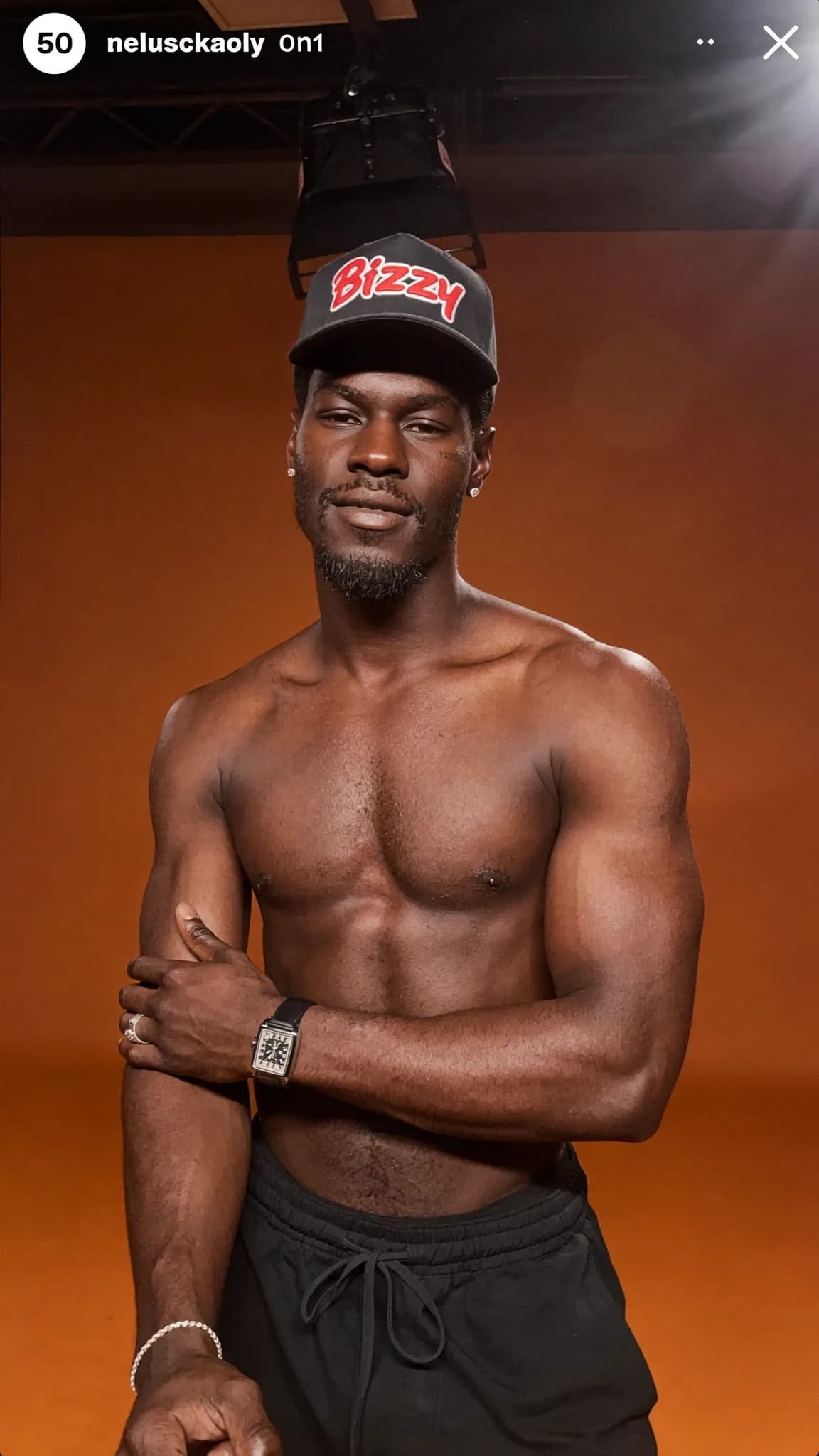 A shirtless man wearing a cap and black shorts poses confidently in a studio setting.