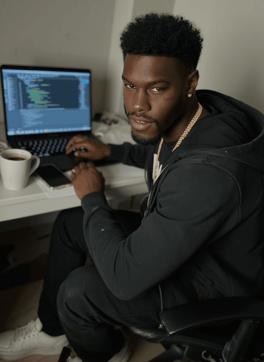 A young man with a pixie cut and septum ring is seated at a desk working on his laptop, wearing casual attire.