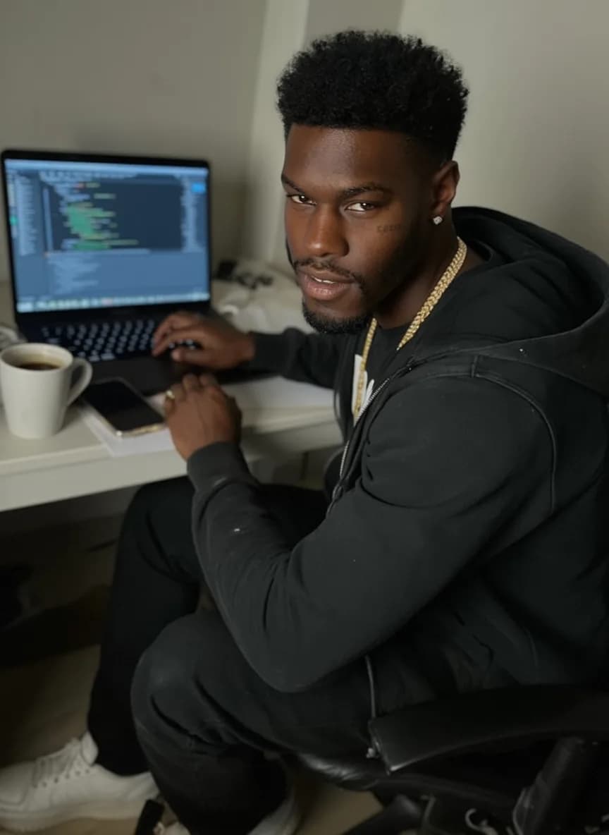 A young man with a pixie cut and septum ring is seated at a desk working on his laptop, wearing casual attire.