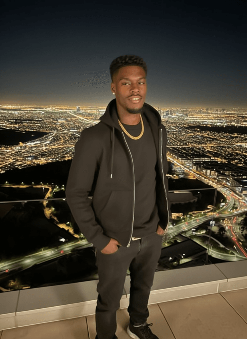 A person stands on a balcony with a cityscape view at night, wearing casual attire and posing for the camera.