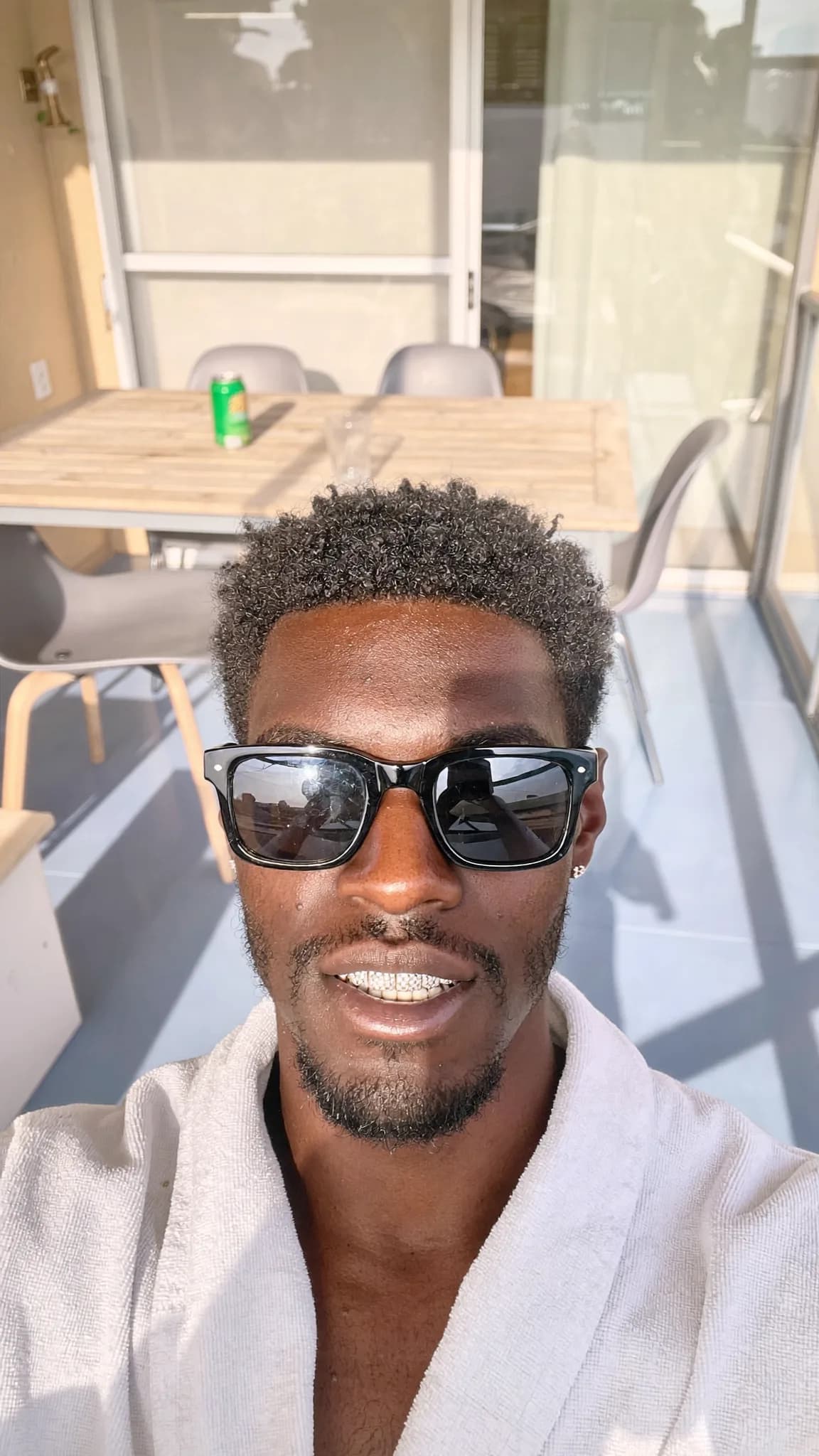 A person wearing sunglasses and a robe poses on a balcony with a table in the background.