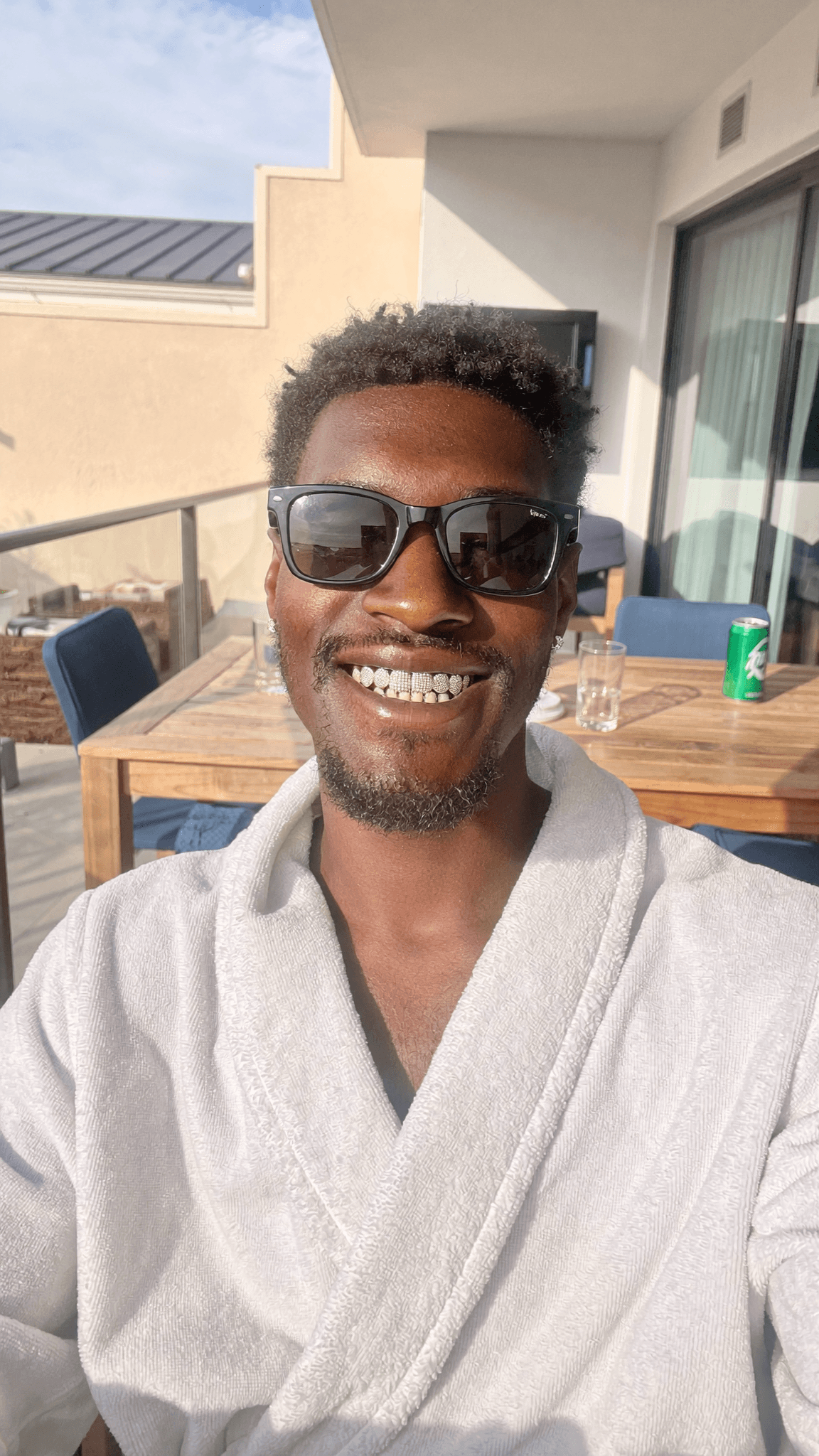 A person wearing a robe and sunglasses is smiling on an outdoor balcony.