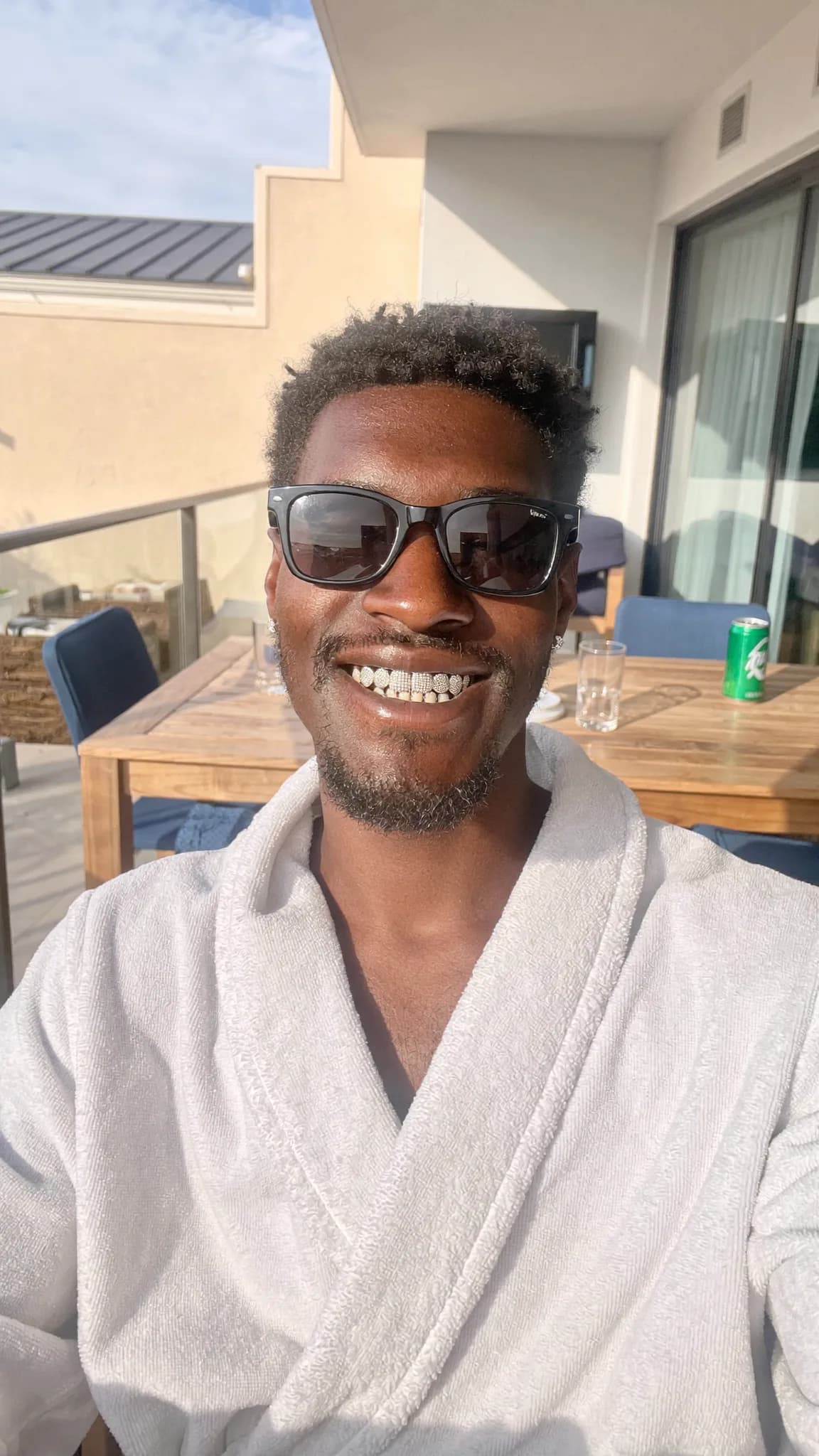 A person wearing a robe and sunglasses is smiling on an outdoor balcony.