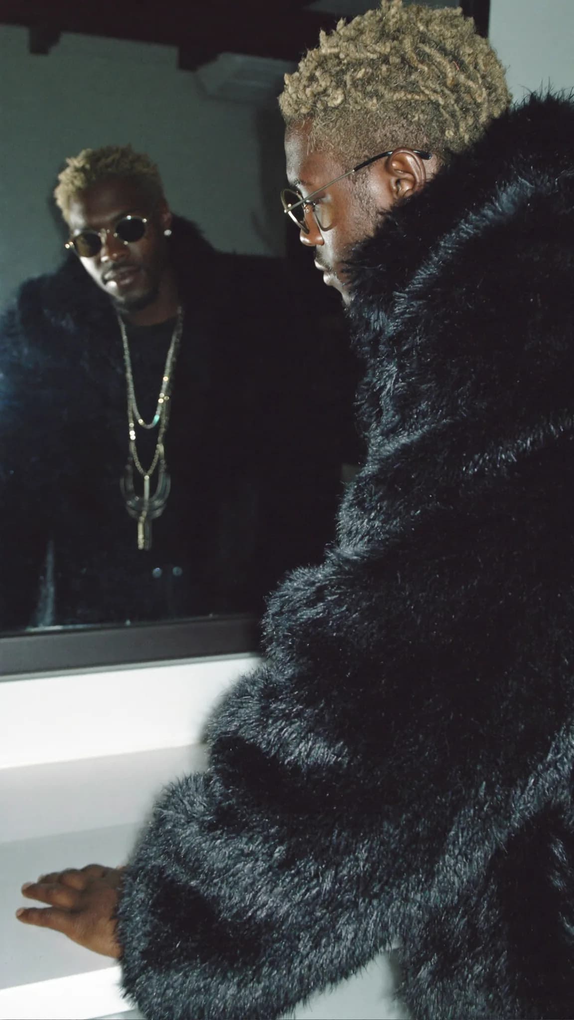 A person in a formal outfit poses confidently with a three-quarter view, wearing sunglasses and jewelry.