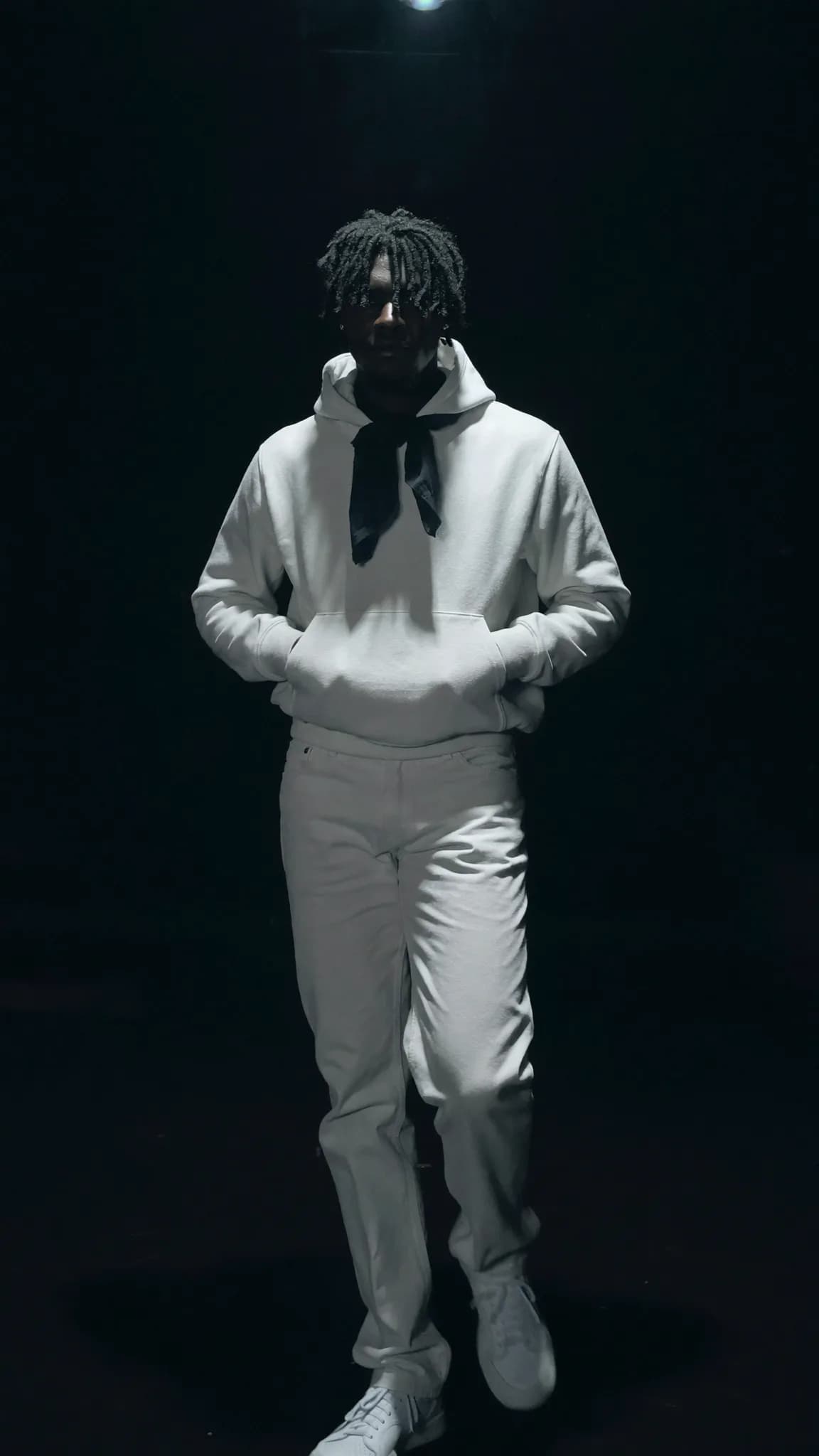 A person in a white outfit stands confidently under focused lighting.