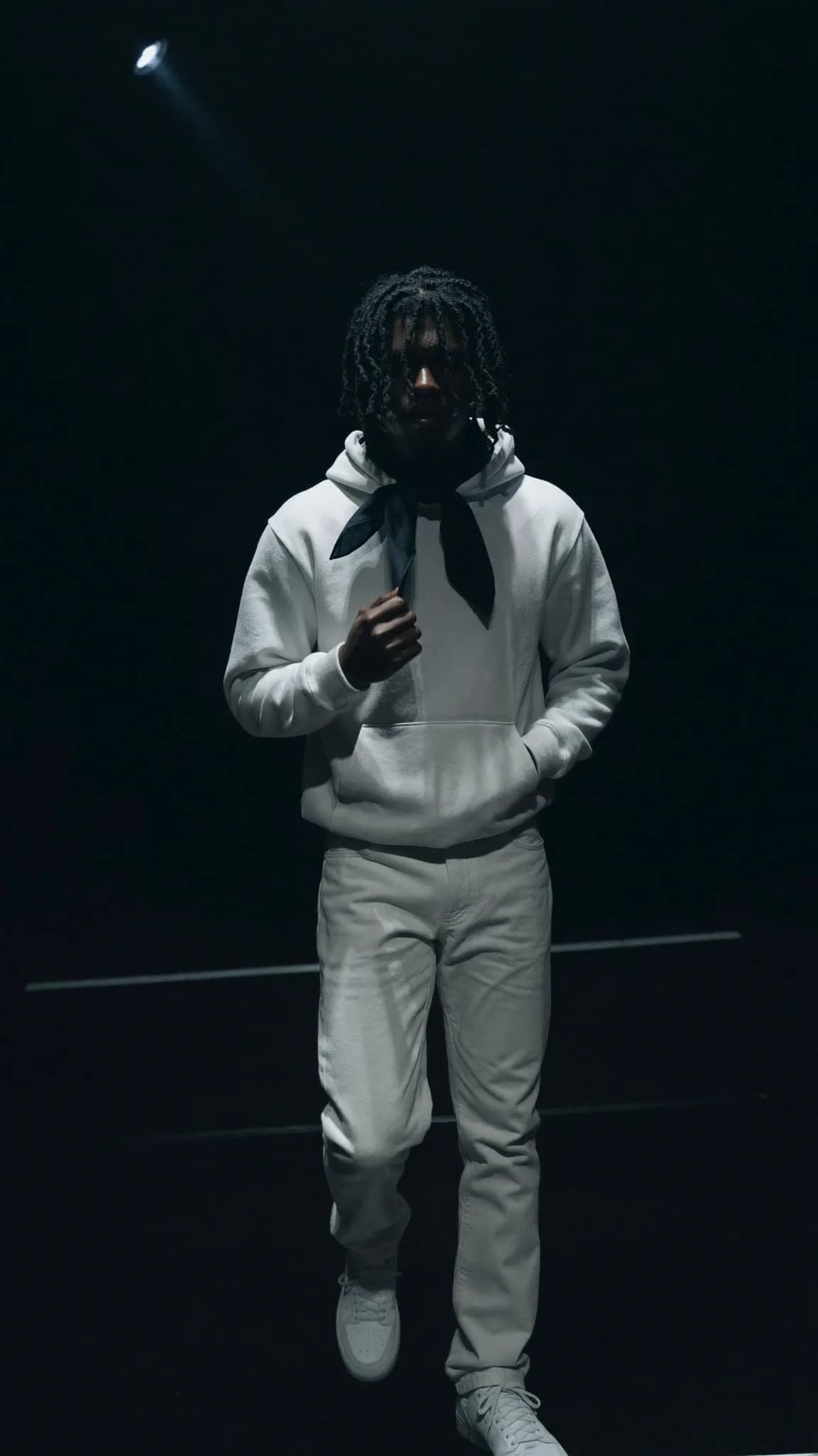 A person in a casual outfit stands under focused lighting on a dark background.