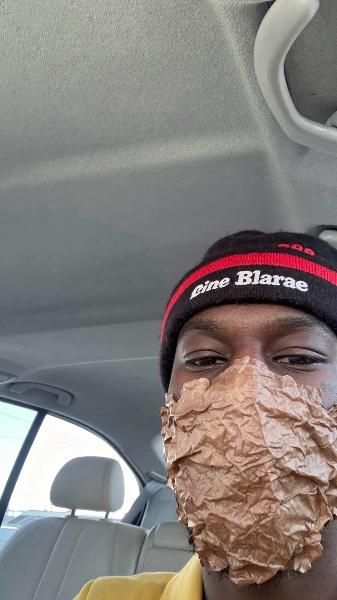 A person wearing a beanie and sitting in a car with a crumpled paper covering their face.