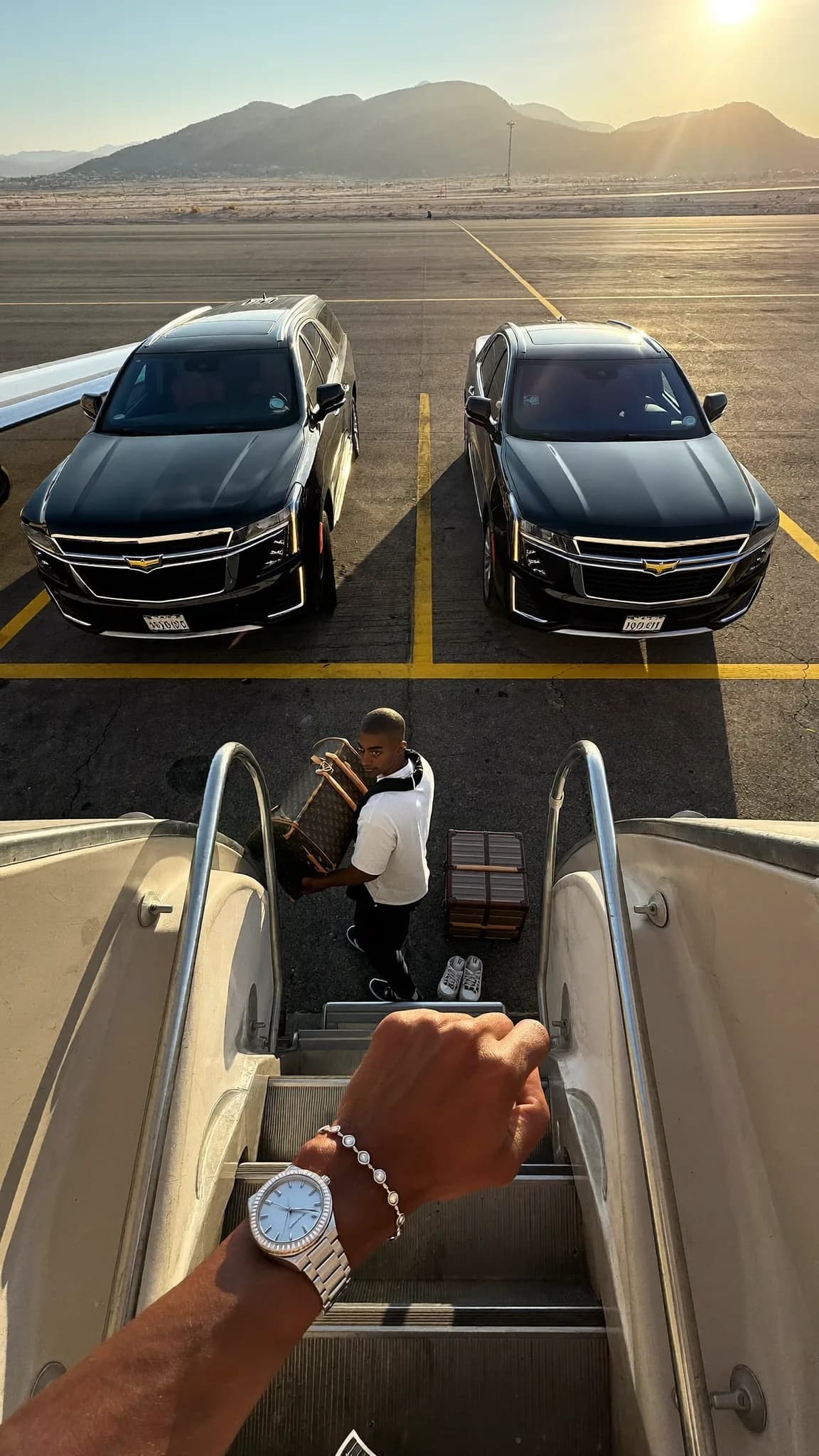 A person wearing an athletic outfit stands on the steps of a private jet, with two luxury cars parked nearby.