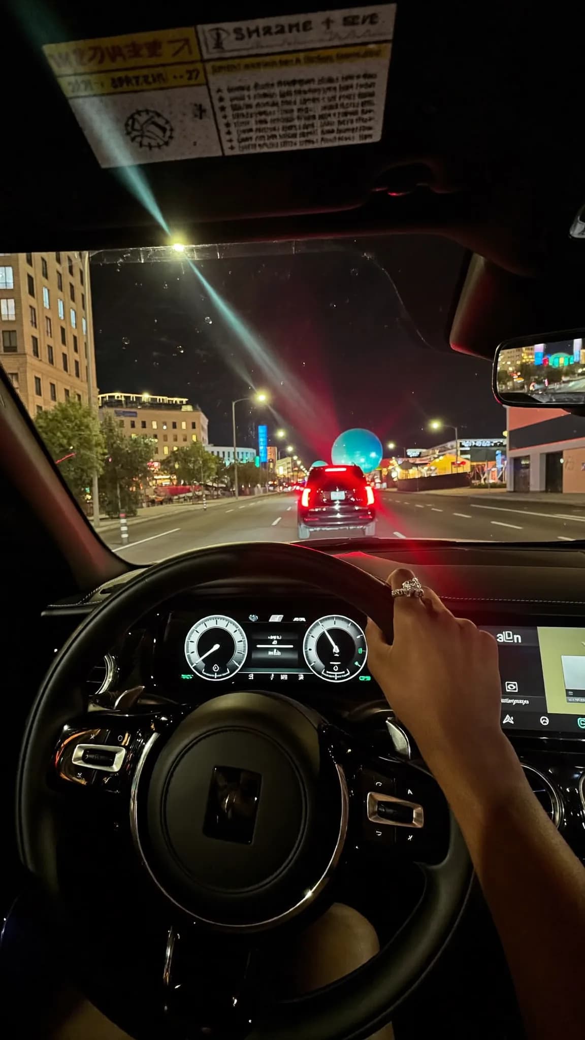 A person drives a car at night with another vehicle ahead, featuring a distinctive blue balloon on the back.