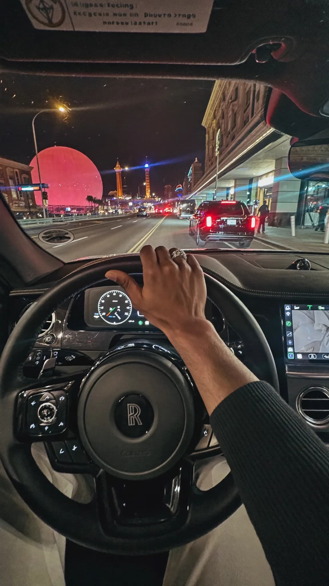 A person drives a luxury car at night with their hand on the steering wheel, passing by brightly lit city landmarks.