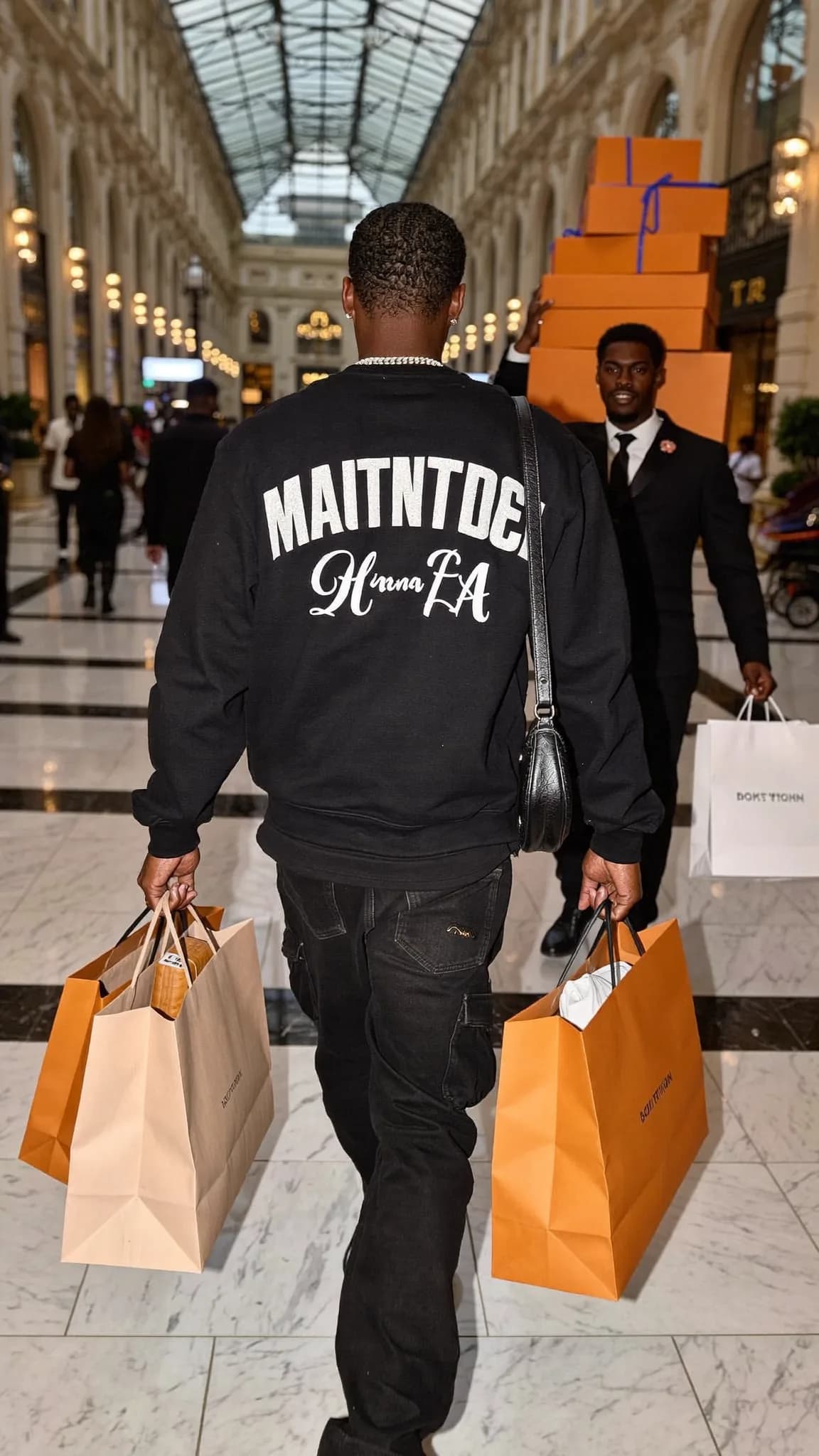 A person in a casual outfit is walking through an indoor space carrying shopping bags, with another individual in the background holding large orange boxes.