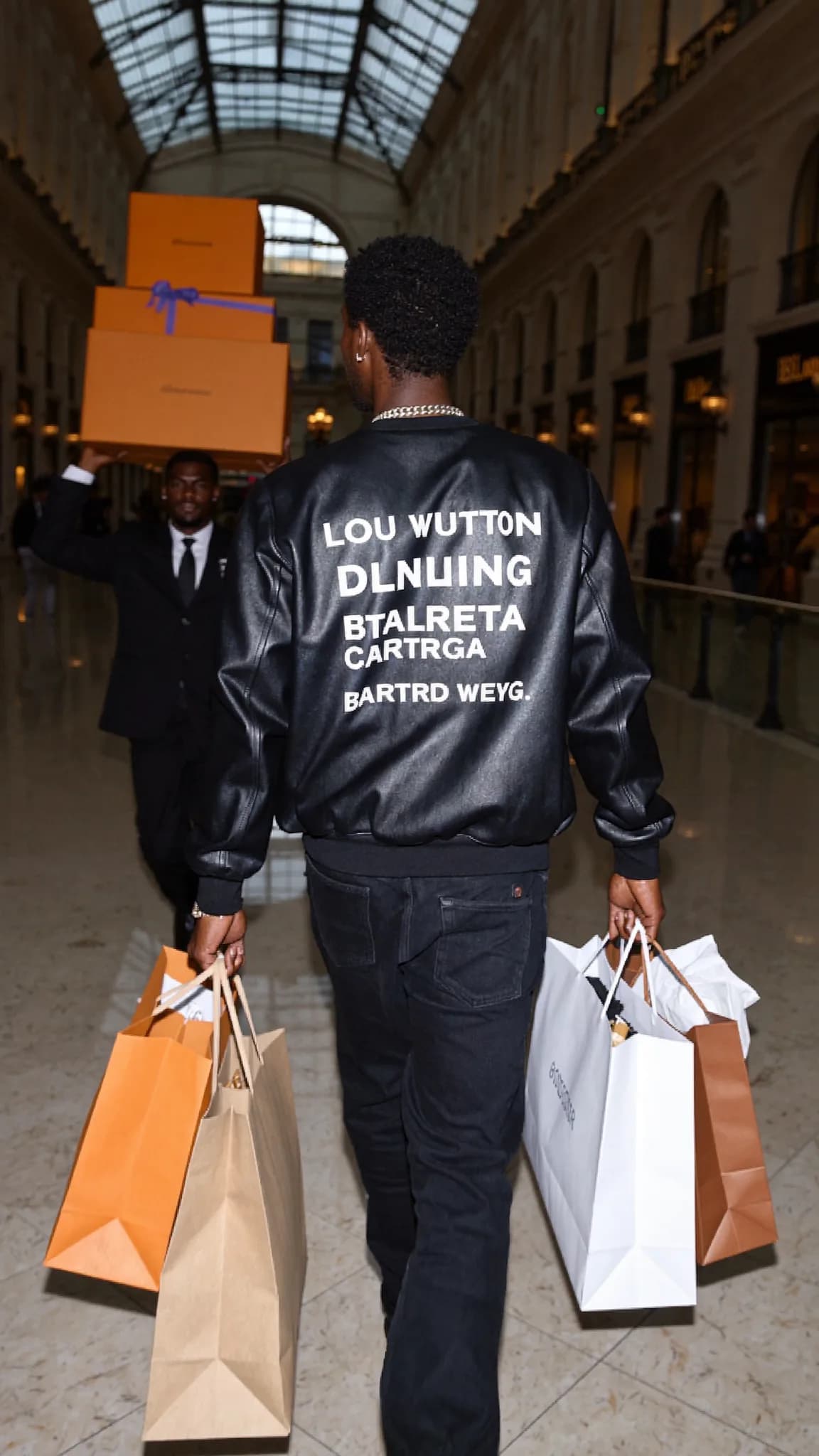 A person in a casual outfit walks down the street carrying shopping bags, with another individual holding large orange boxes behind them.