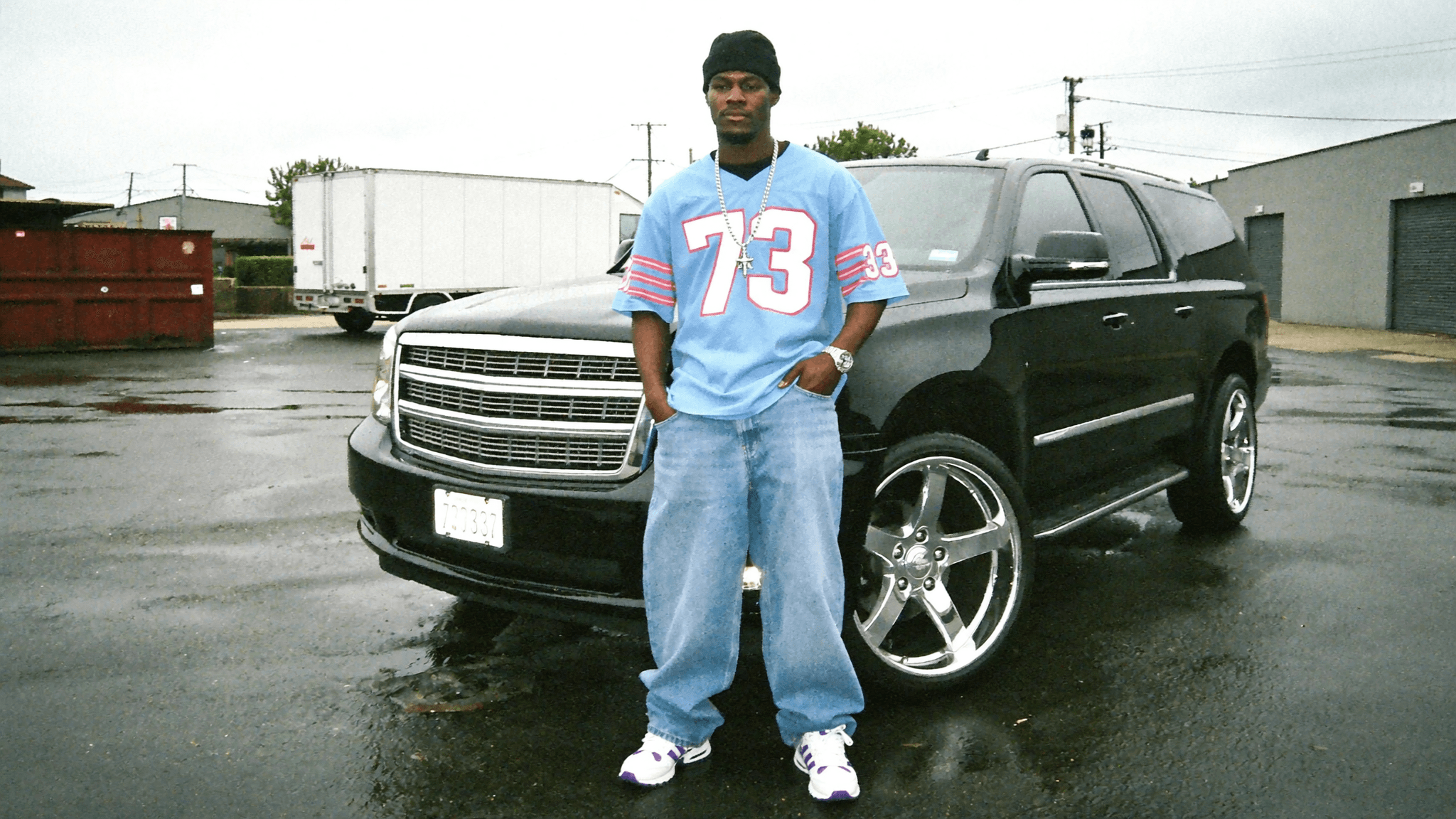 A man stands in front of a black SUV wearing casual clothing and posing outdoors.