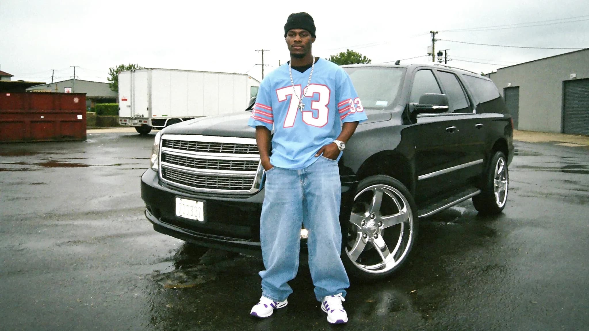 A man stands in front of a black SUV wearing casual clothing and posing outdoors.