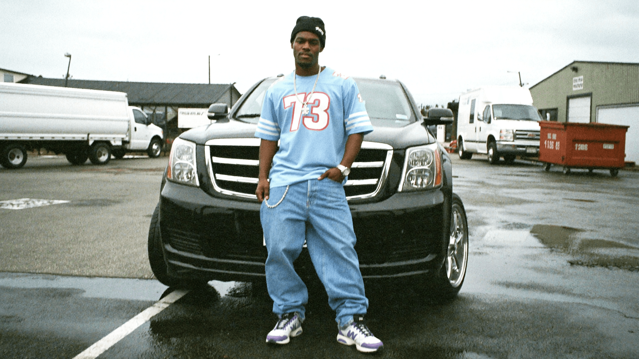 A person stands in front of a black car wearing casual clothing and posing outdoors.