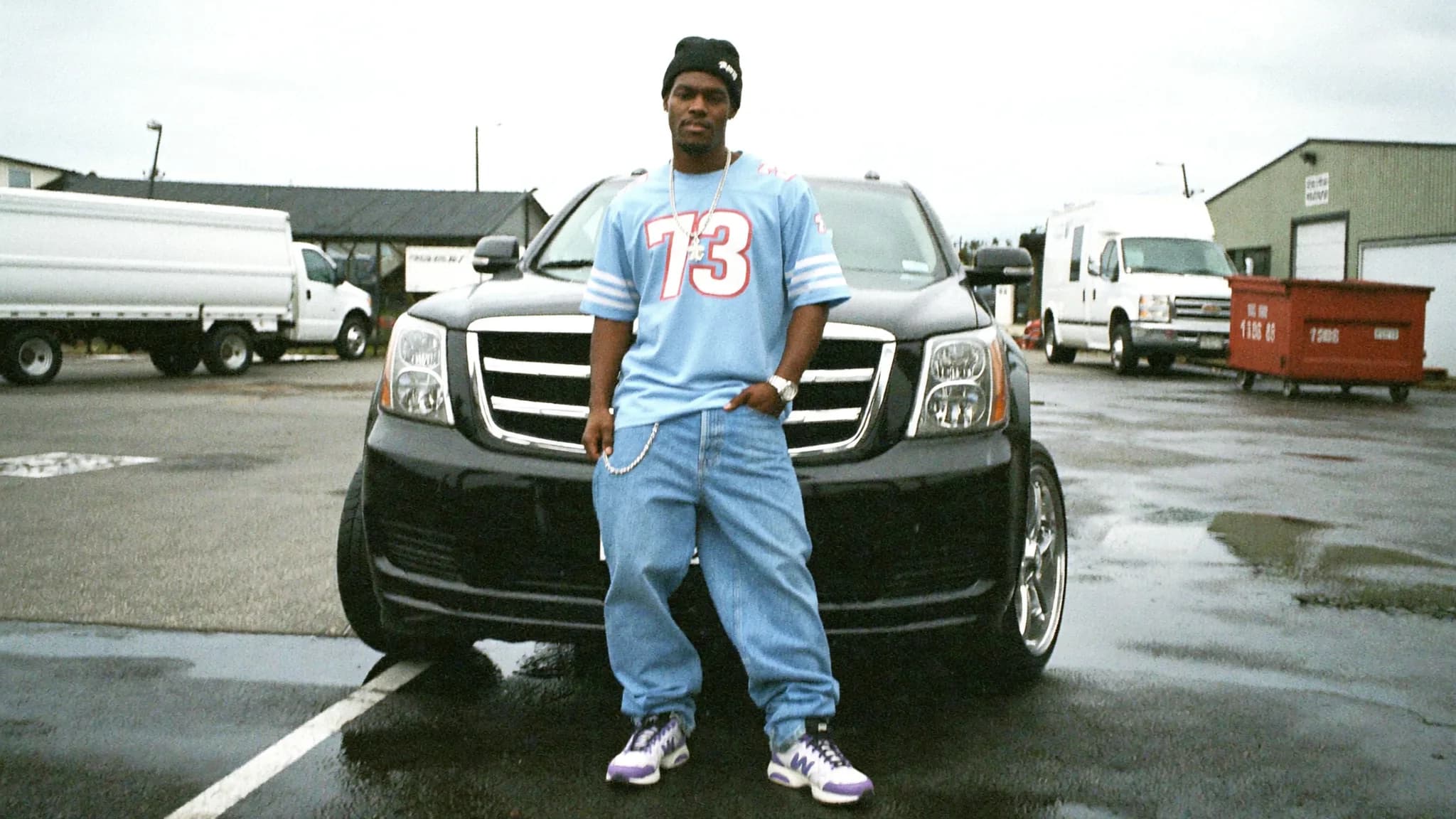 A person stands in front of a black car wearing casual clothing and posing outdoors.