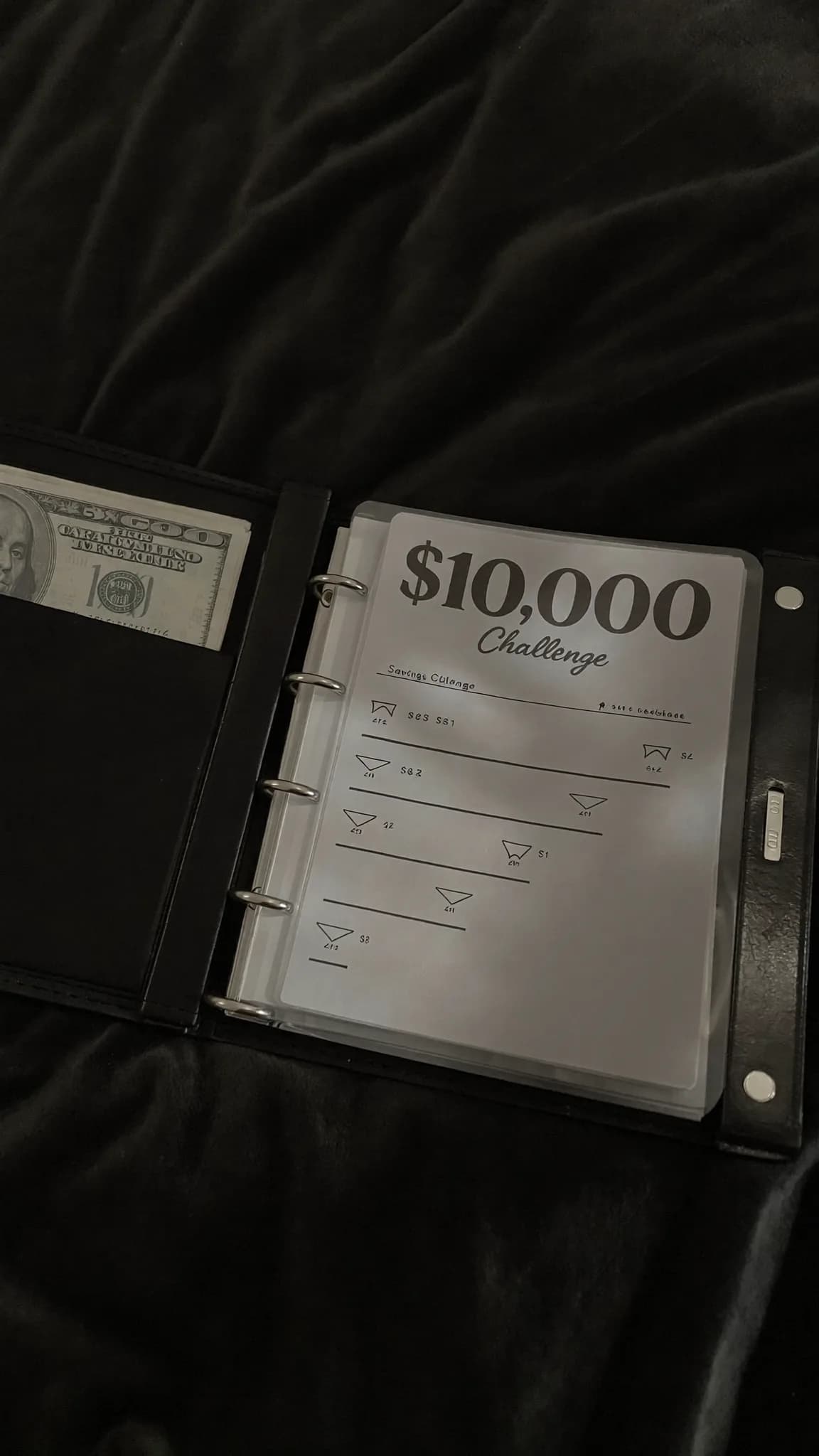 A binder with a $10,000 challenge and savings categories is placed on a bed.