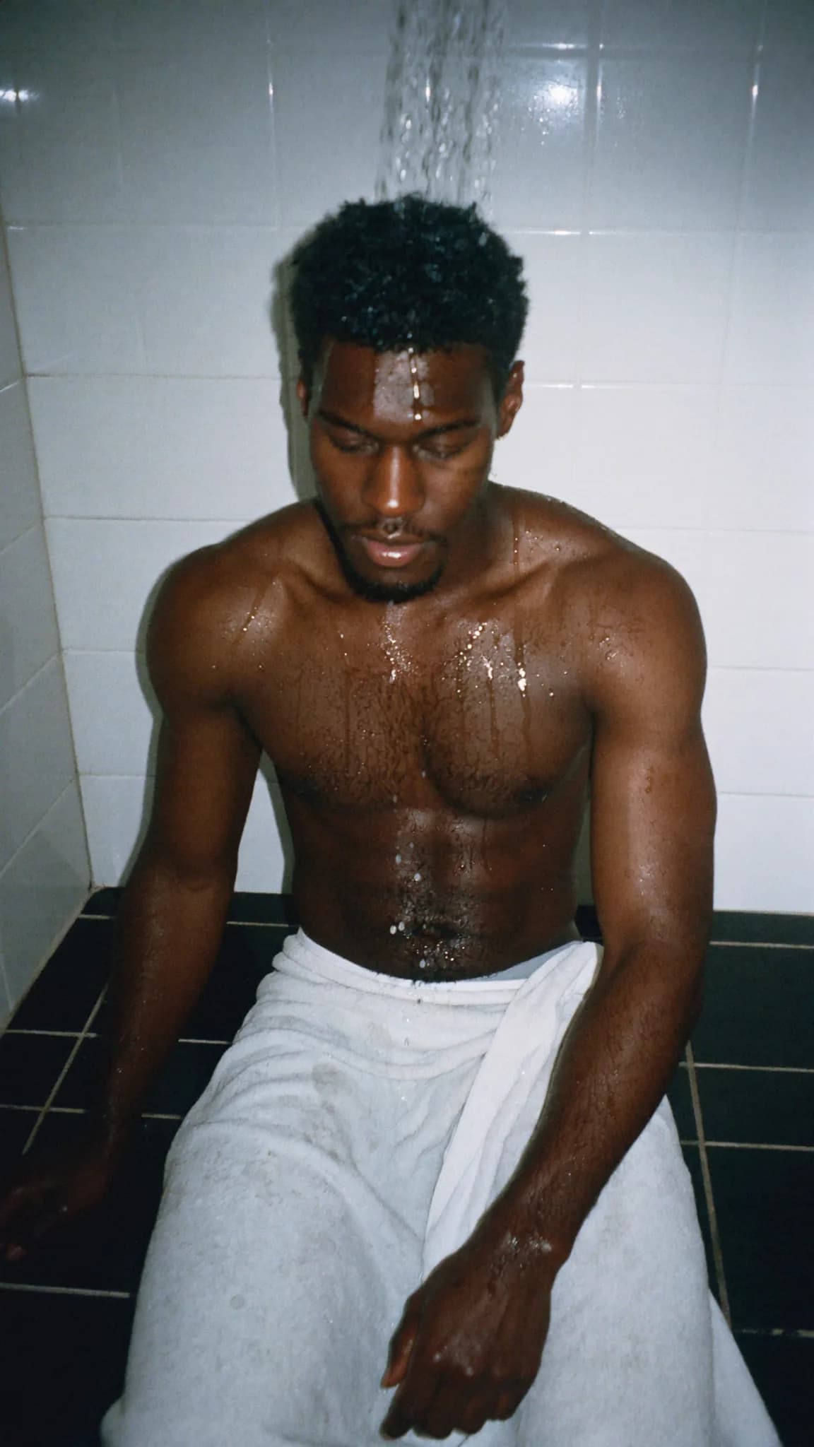 A person is seated in a bathroom, wet and wrapped in a towel.