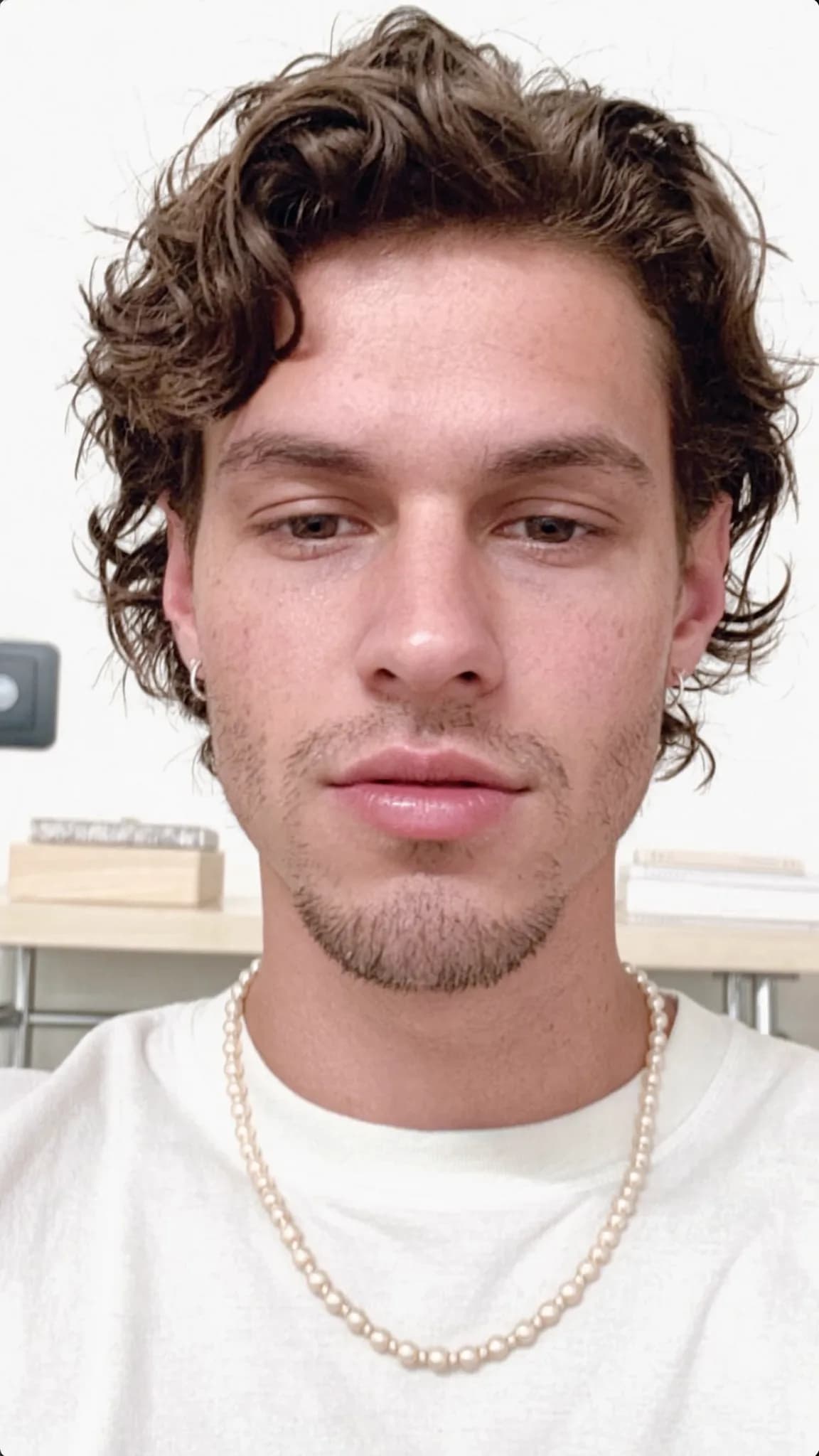 A young man with curly hair and a pearl necklace poses in a studio setting.