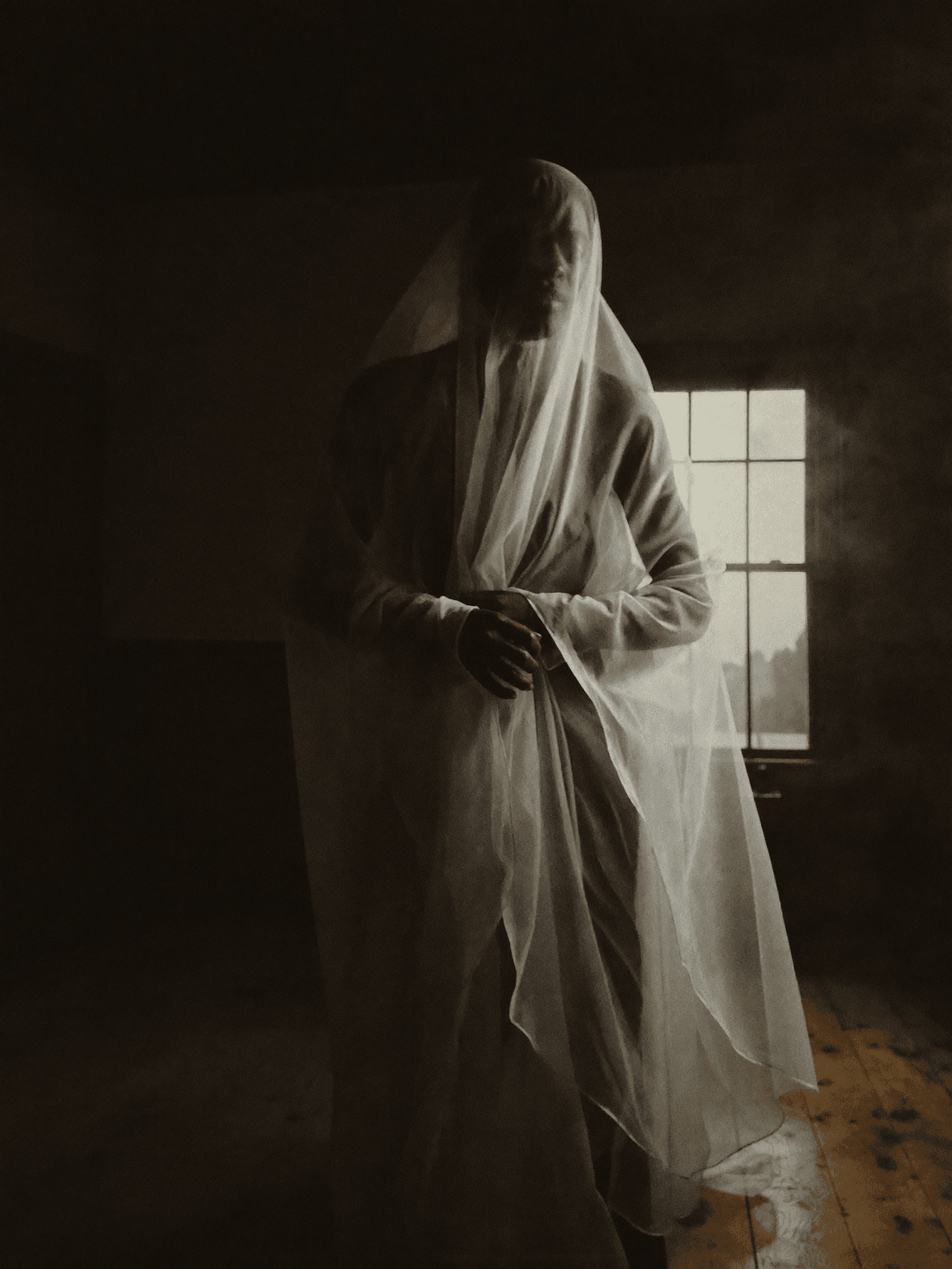 A figure draped in a sheer fabric stands in a dimly lit bedroom, exuding an ethereal and intimate atmosphere.