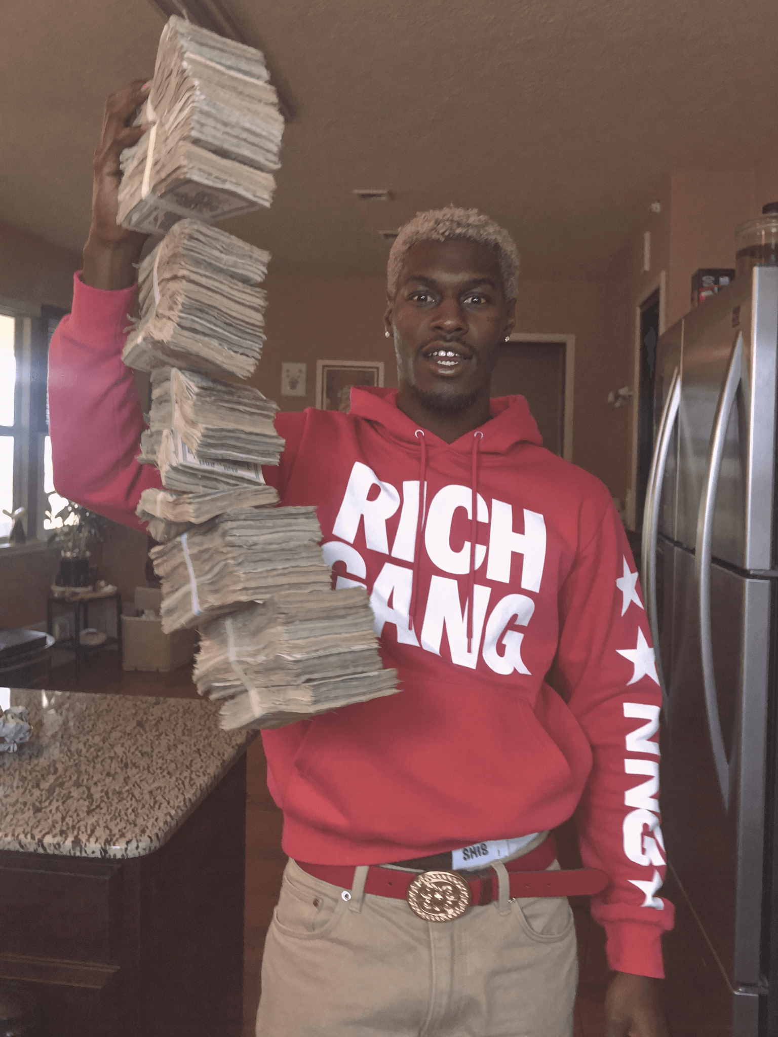 A person in a red hoodie holds stacks of cash while standing in a kitchen.