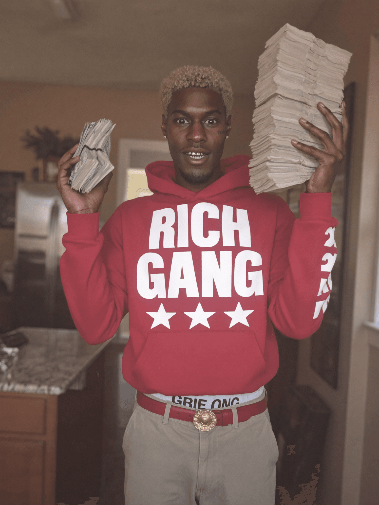 A person in a red hoodie labeled 'RICH GANG' holds stacks of money and papers, standing confidently in a kitchen.