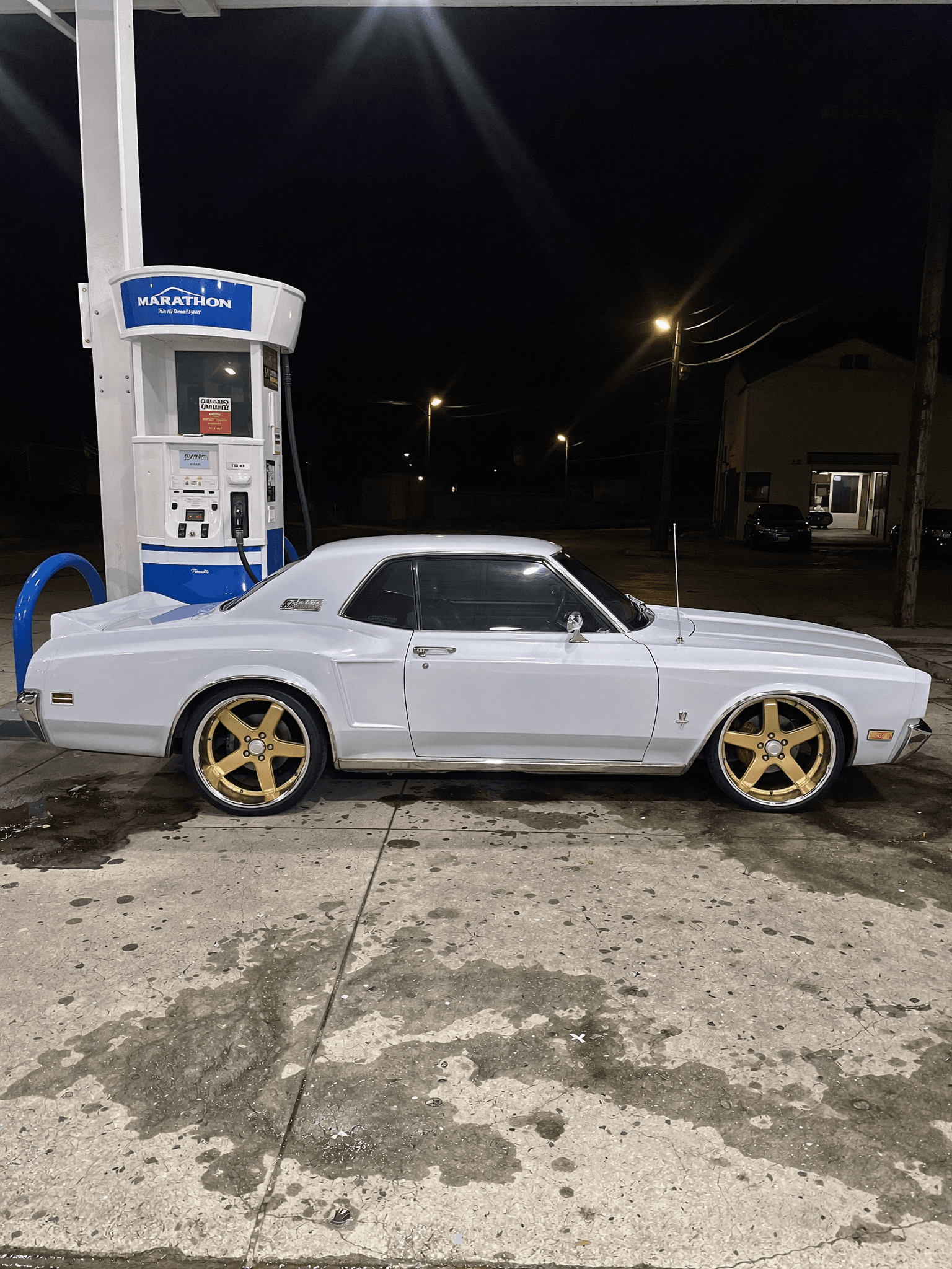 A white classic car with gold rims is parked at a gas station during the night.