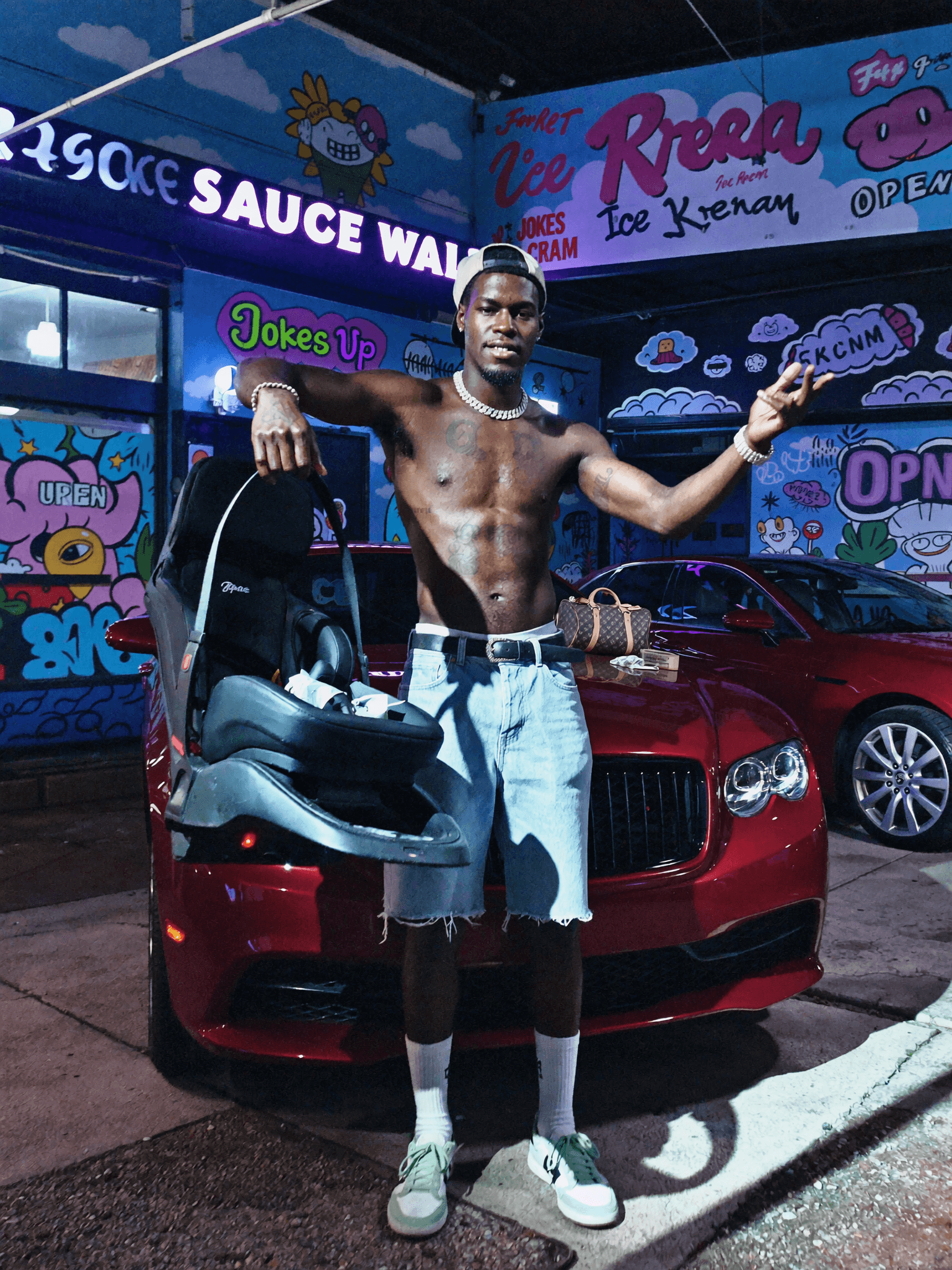 A person stands confidently in front of a red car, wearing casual athletic attire and posing with one hand on the vehicle's hood.