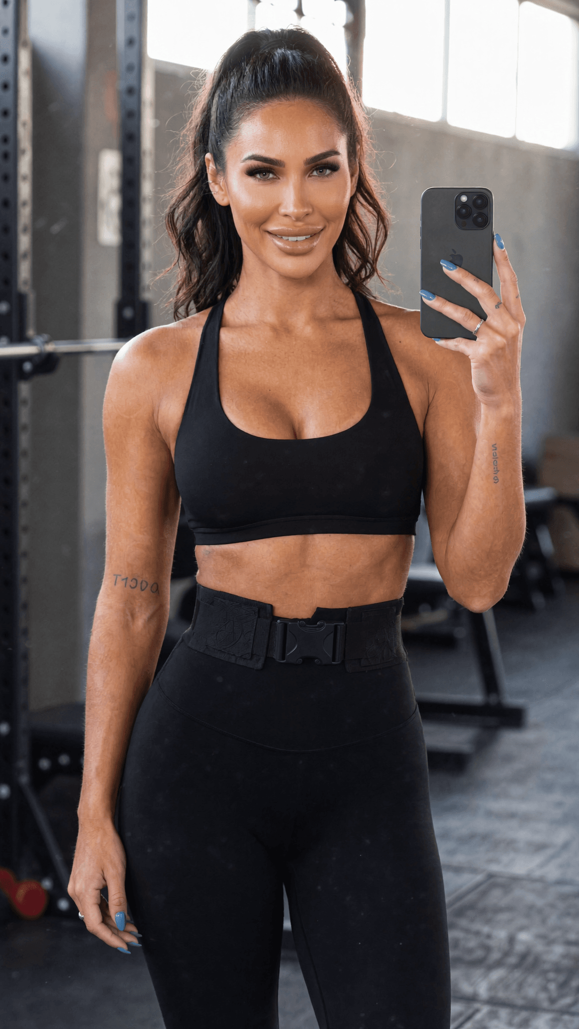 A woman in a black sports bra and leggings poses confidently in a studio, holding her phone.