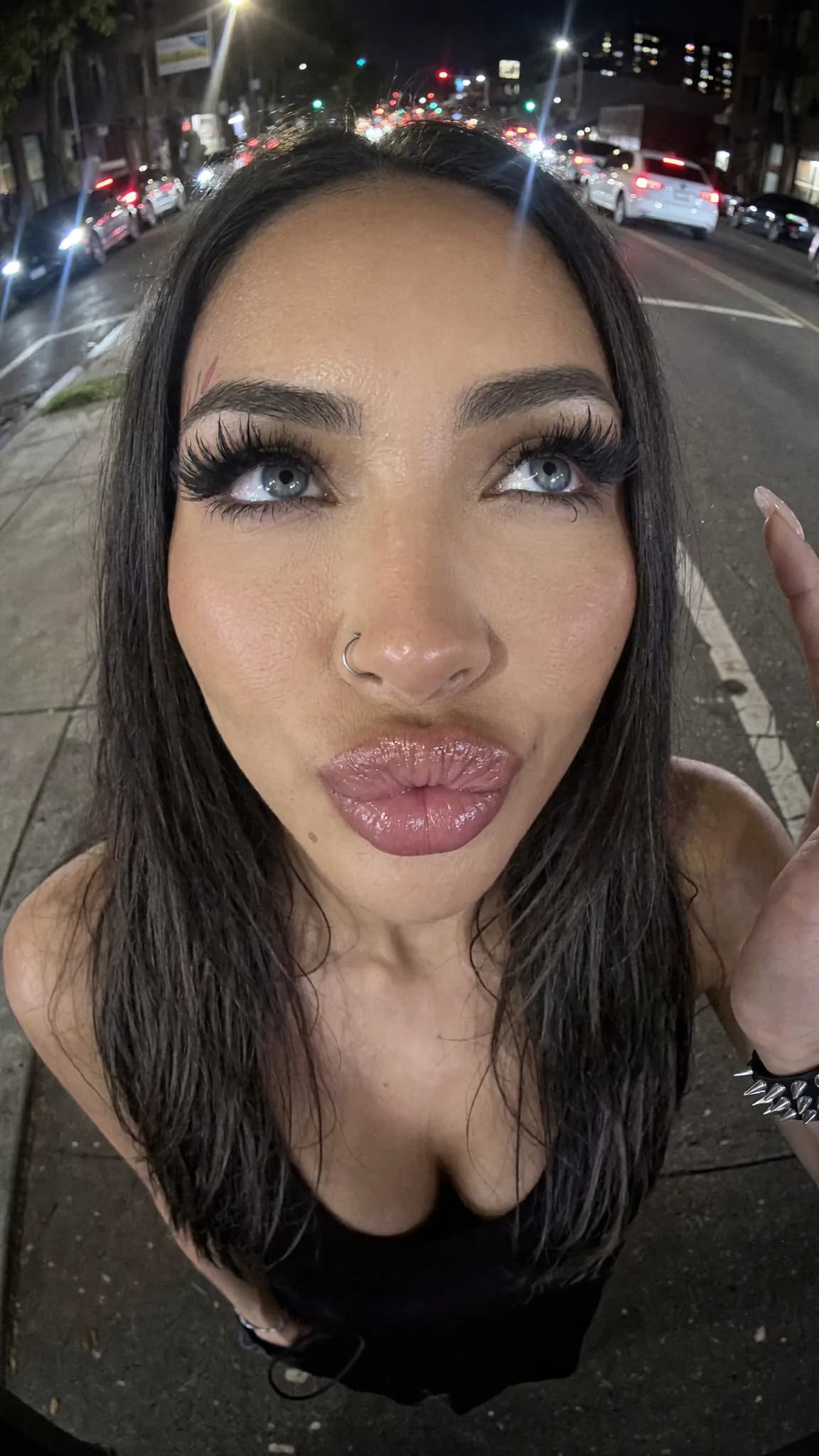 A woman with long black hair and a septum ring poses on the street at night, wearing casual clothing.