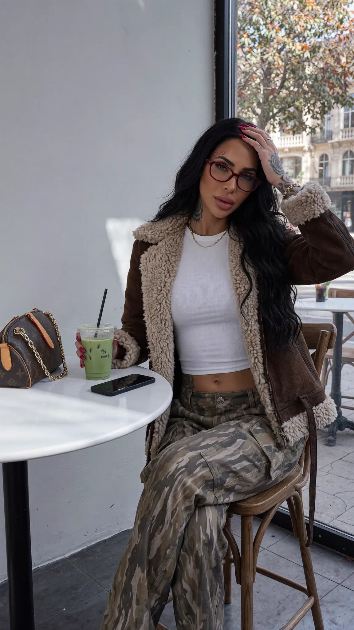 A woman with long black hair and glasses sits at an outdoor table, holding a green drink.