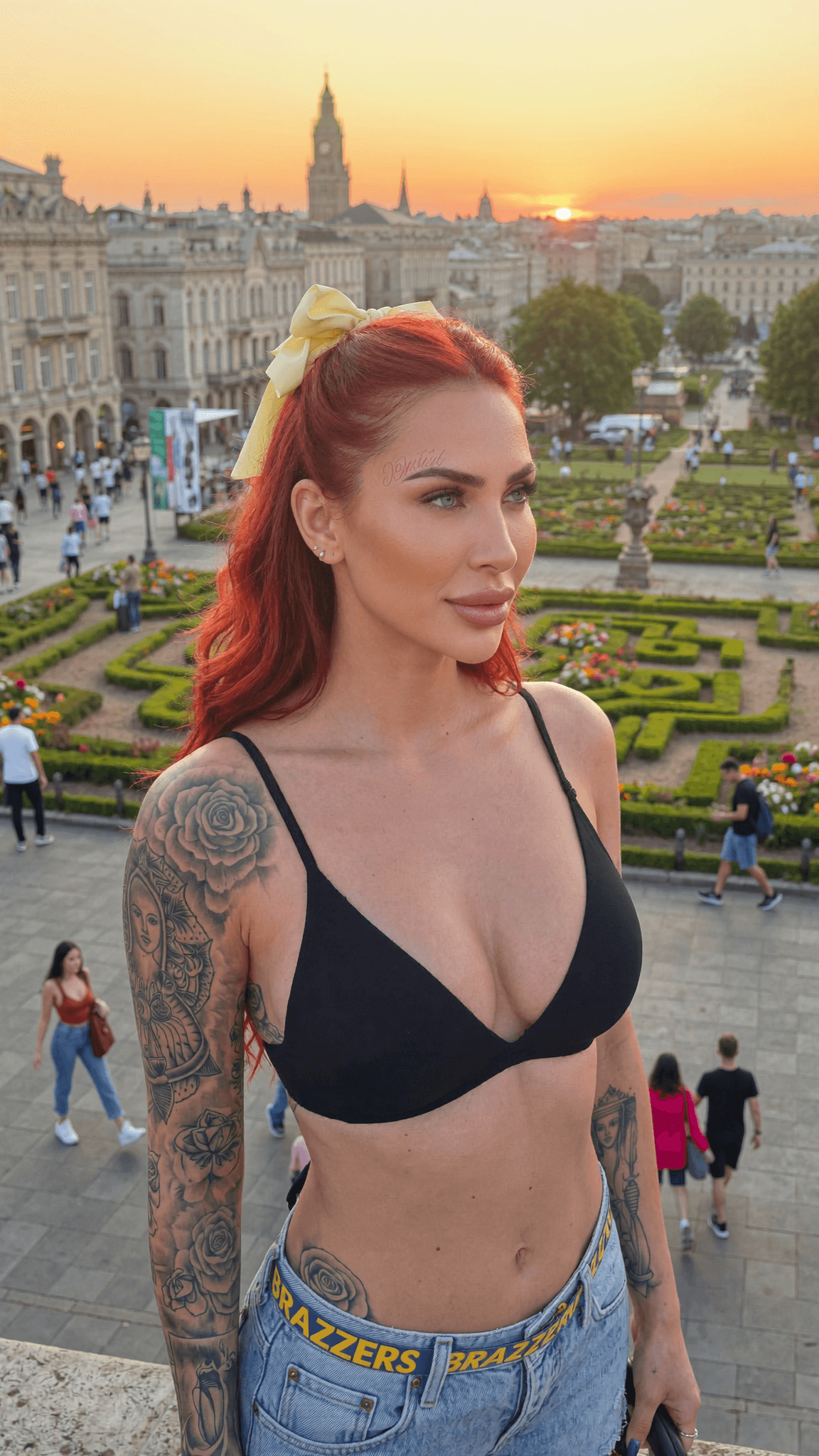 A woman with red hair and tattoos stands in a scenic outdoor setting during sunset, wearing a swimsuit.