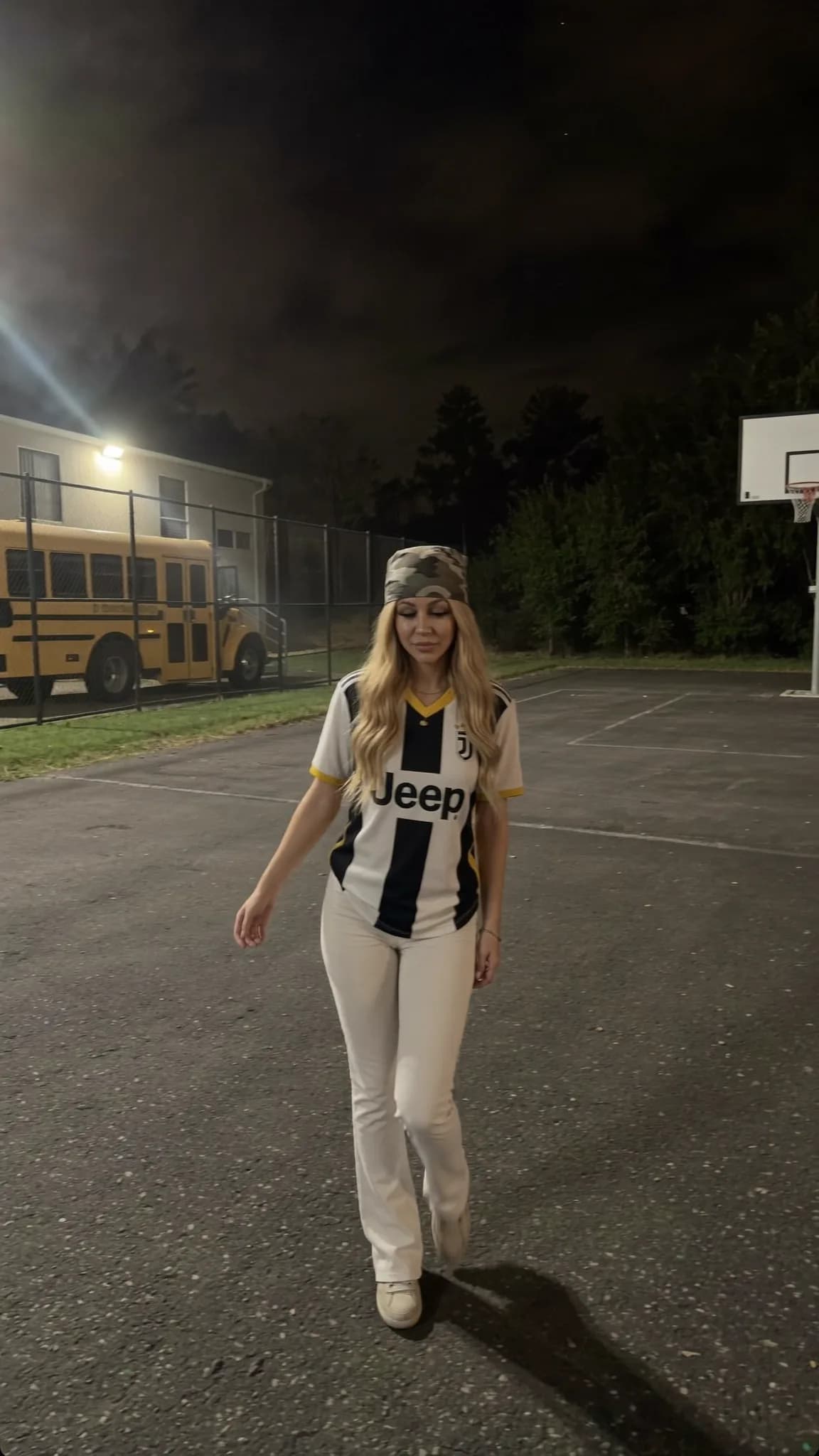 A woman in a sports jersey and camouflage headband stands on an outdoor basketball court at night.