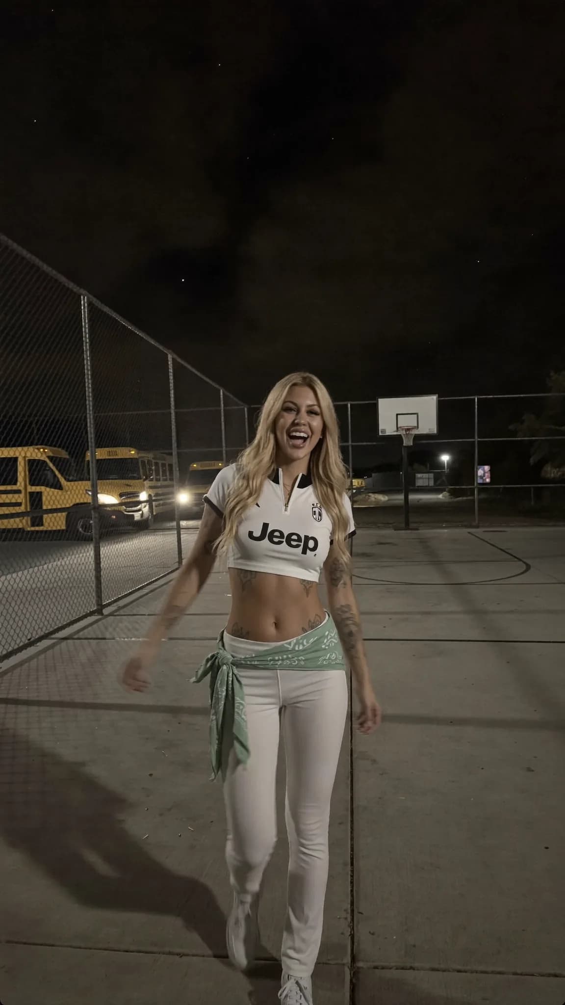 A woman in a crop top and leggings stands on an outdoor basketball court at night, smiling.