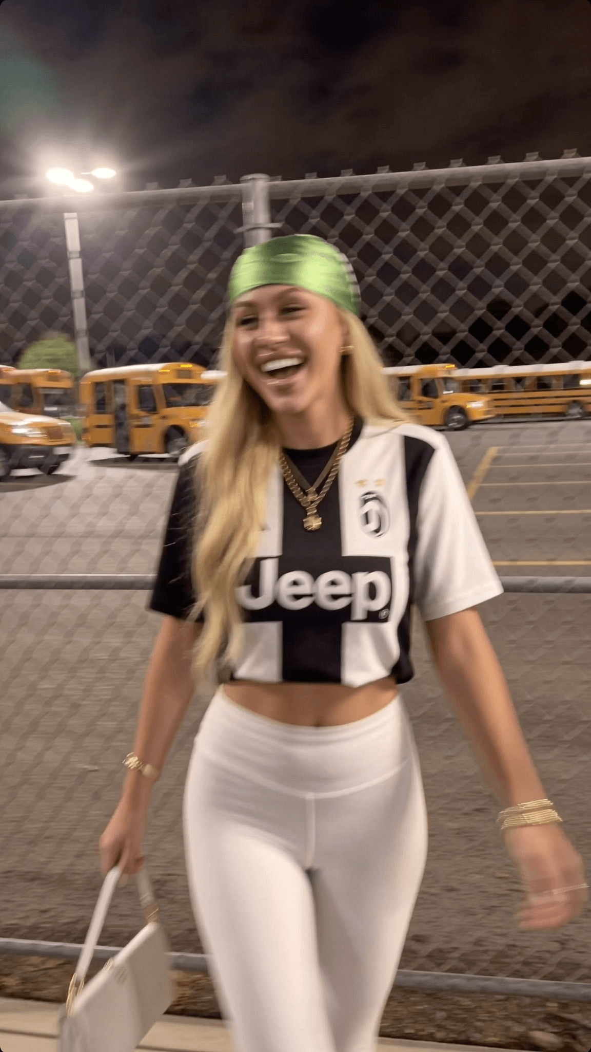 A woman in a Juventus jersey and white leggings is walking outdoors at night, smiling.
