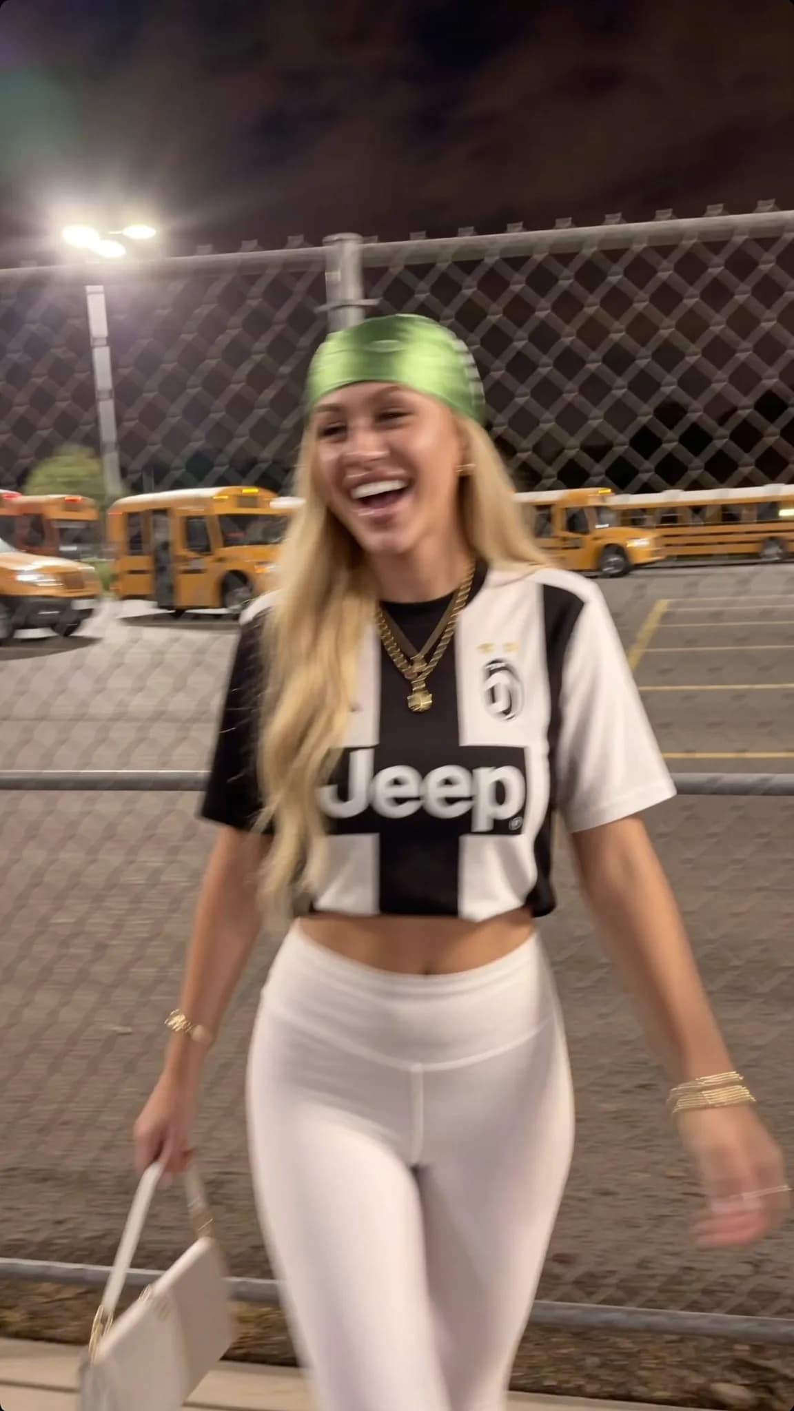 A woman in a Juventus jersey and white leggings is walking outdoors at night, smiling.
