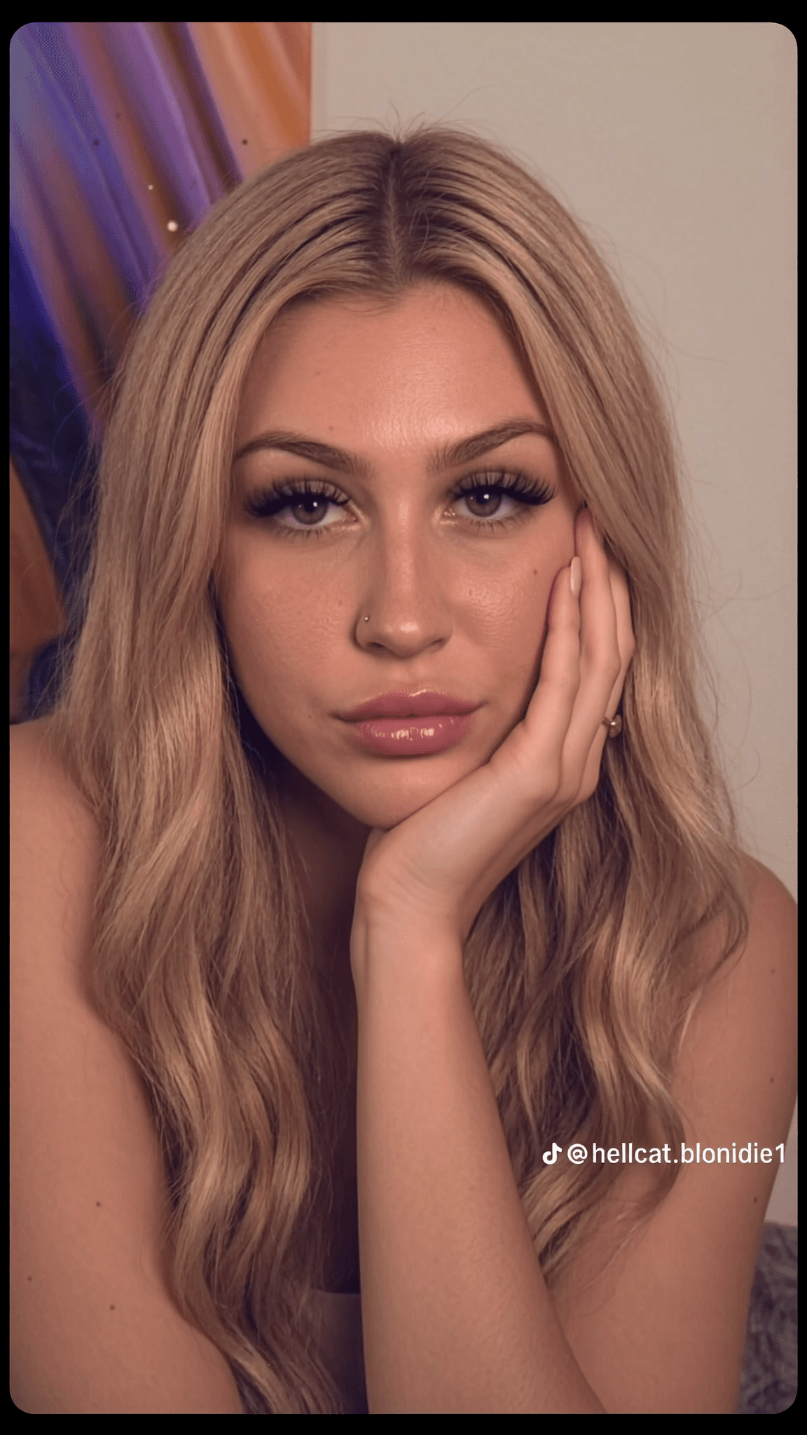 A woman with long blonde hair poses in a studio setting, wearing minimal makeup and jewelry.